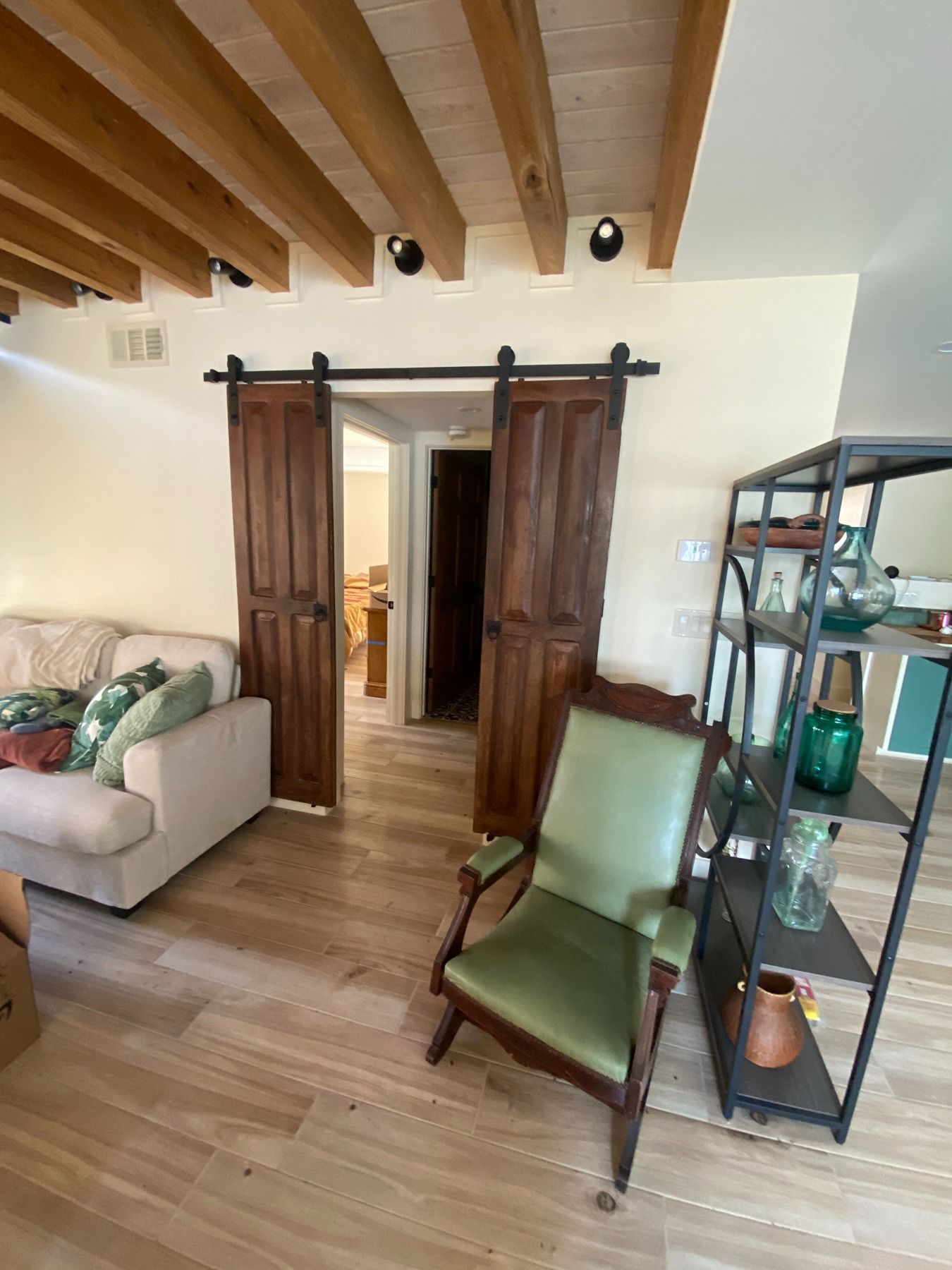 A modern living room with wood ceiling beams, double sliding barn doors, a sofa, a green chair, and a metal shelf.