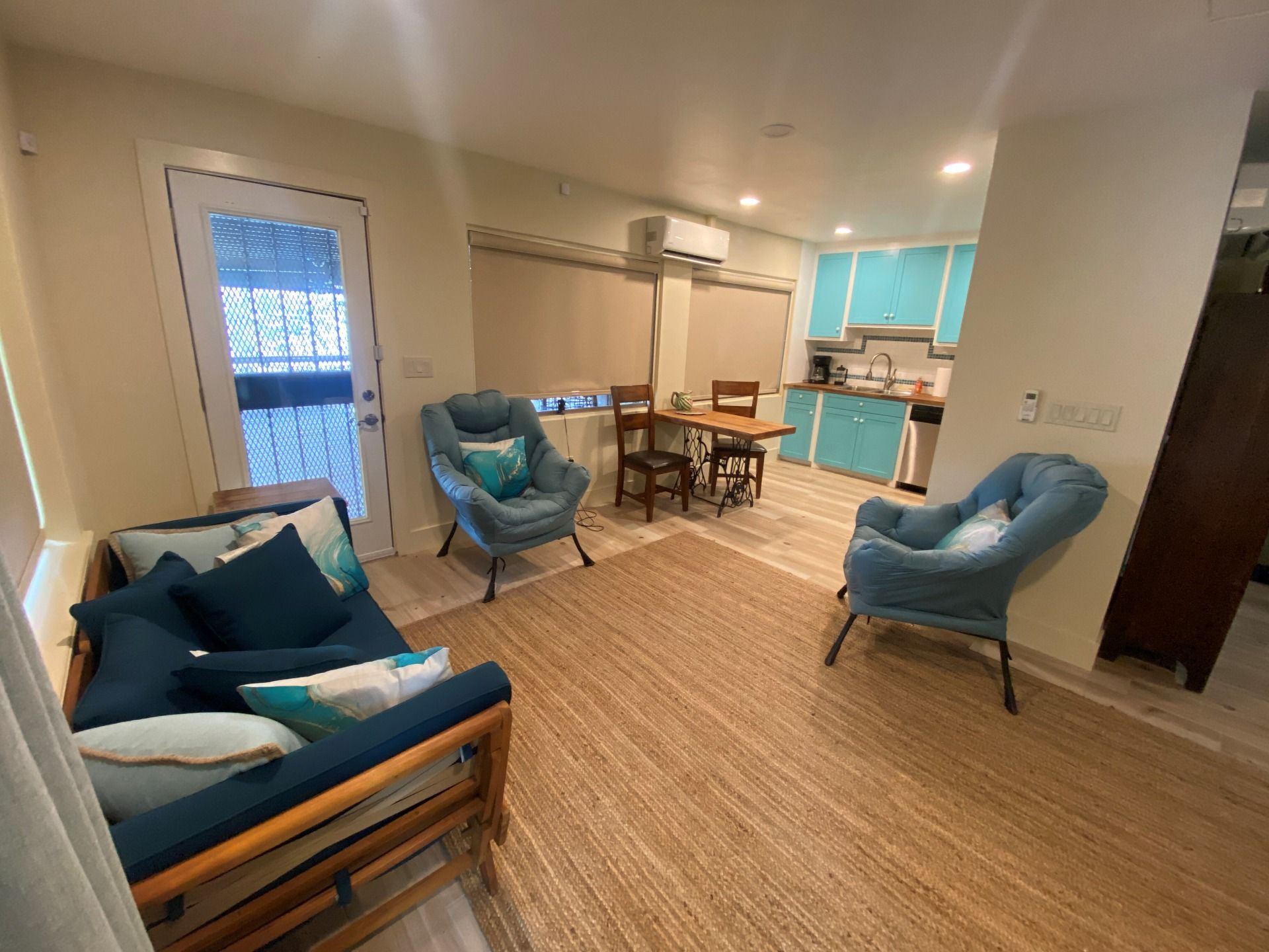 A furnished living space with a blue sofa, two matching armchairs, and a small dining set next to a turquoise kitchen.