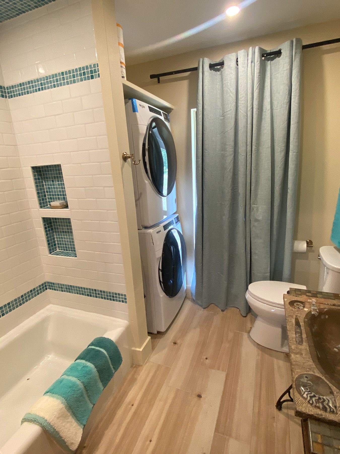 A bathroom with light wood floors, a white tub, stacked washer-dryer, light blue curtains, and a stone-textured vanity.