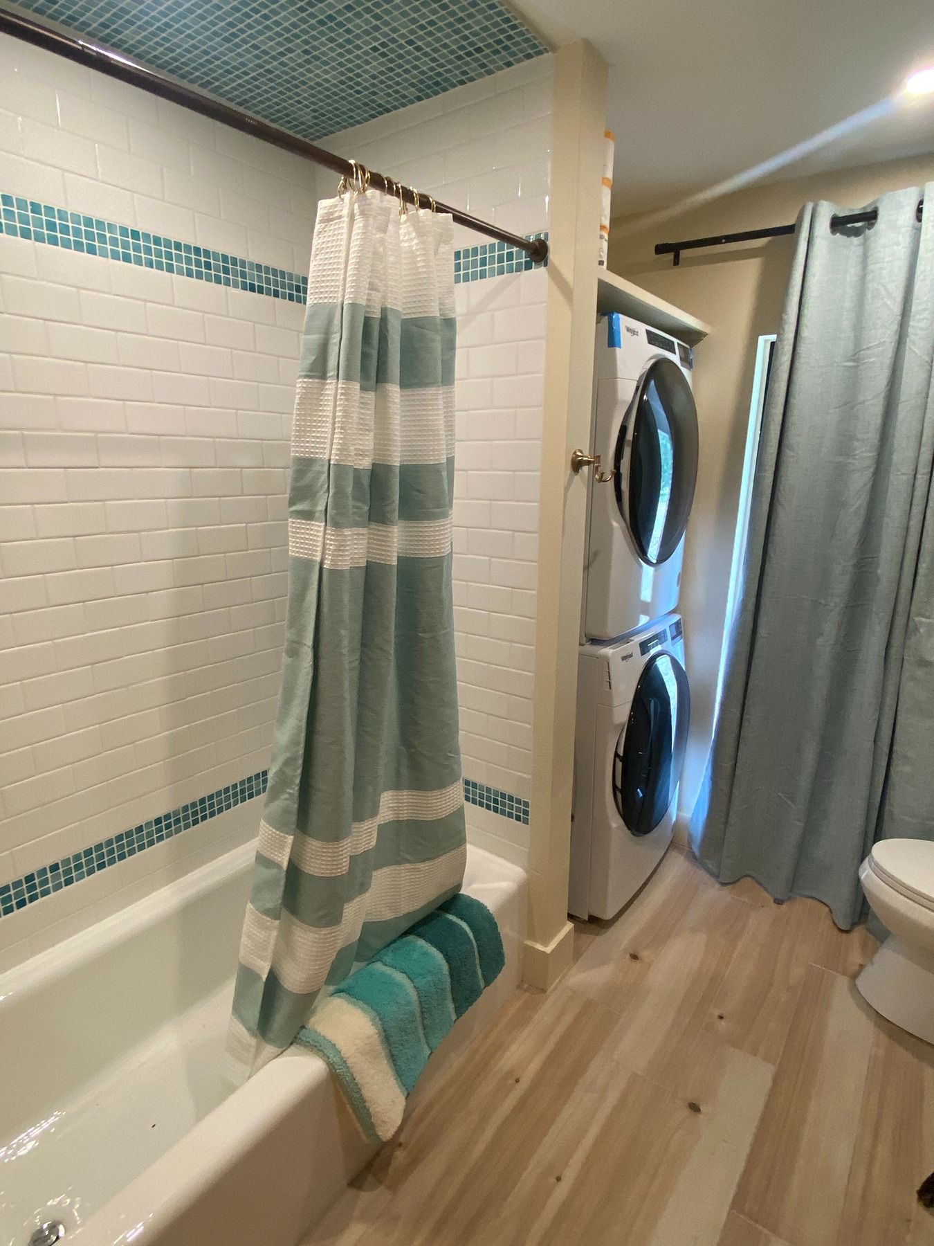 A bathroom with a white tiled tub, blue striped shower curtain, stacked washer-dryer, wood floor, and blue curtain.