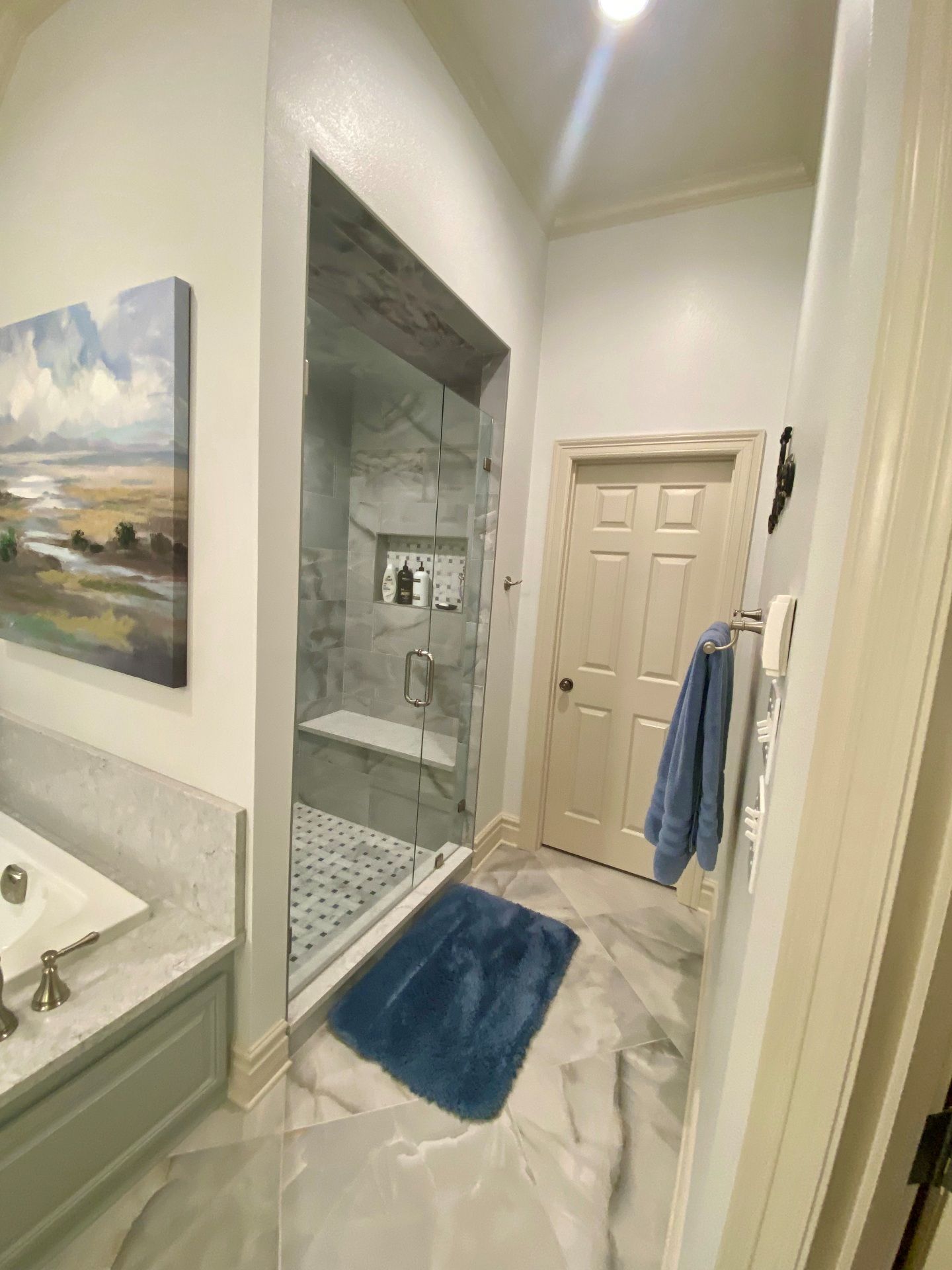 A view of a bathroom showing a tiled walk-in shower with a bench, a blue bath mat, a doorway, and framed wall art.