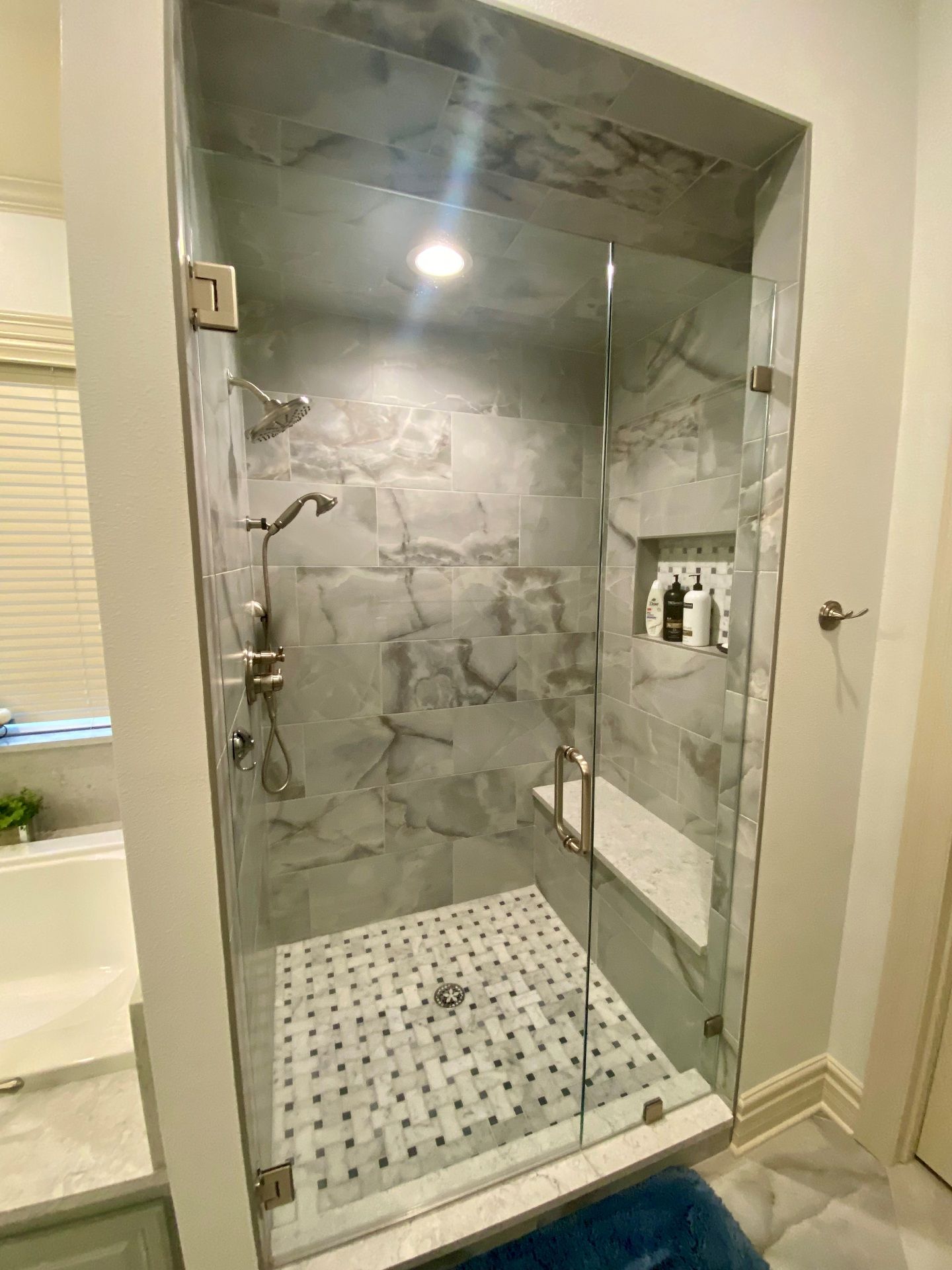 A walk-in shower with gray marbled tiles, a built-in bench, a glass door, and a white and gray mosaic-tiled floor.