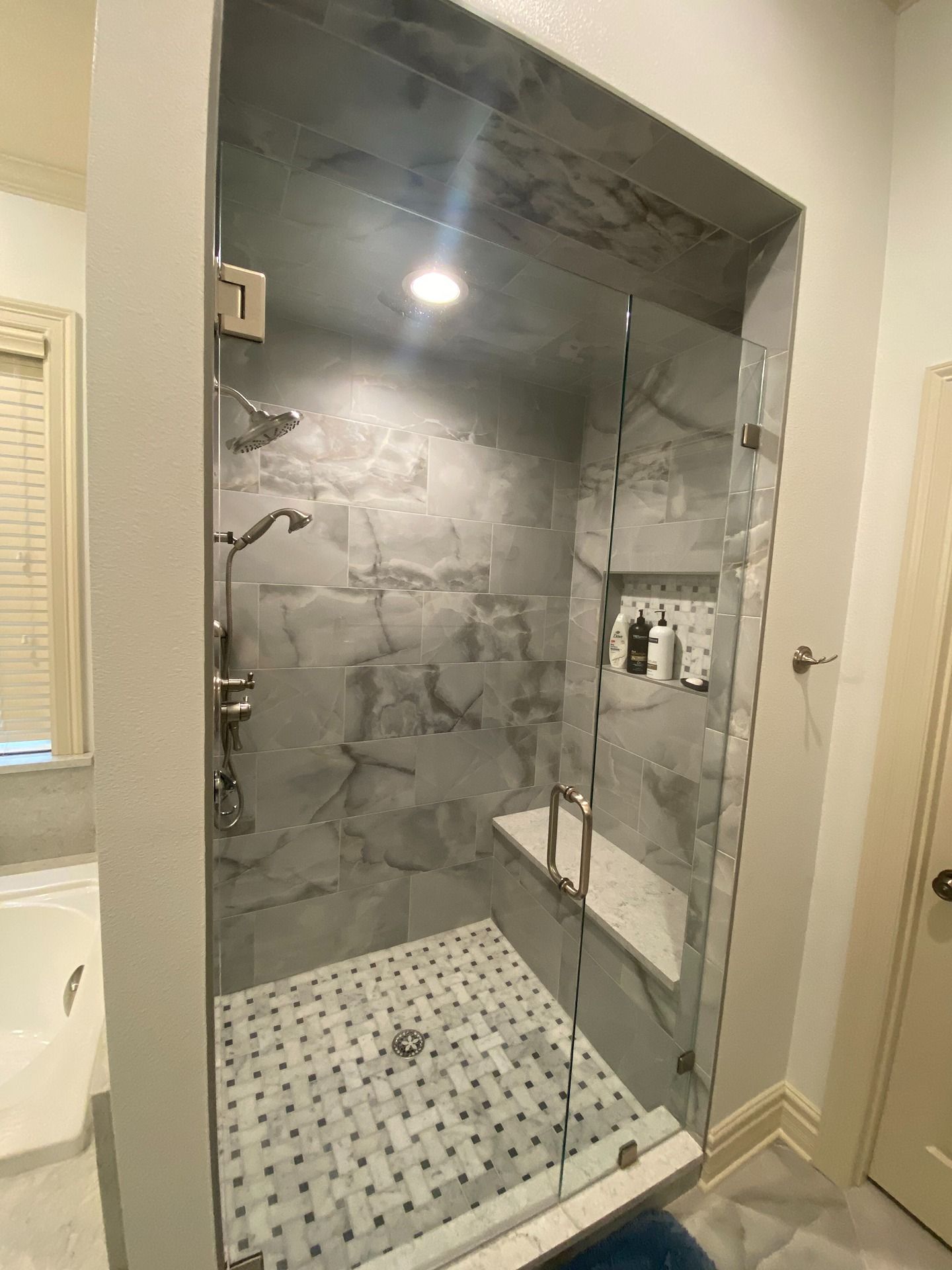 A walk-in shower with gray tiled walls, a marble bench, glass door, and a patterned tile floor in a modern bathroom.