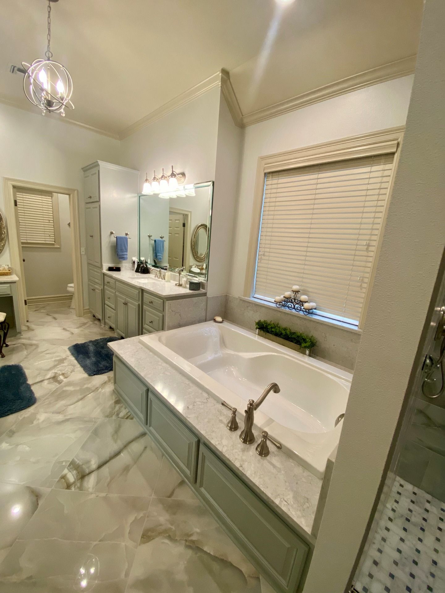 A bright, modern bathroom featuring a white soaking tub with a marble surround, grey cabinetry, and light tile floors.