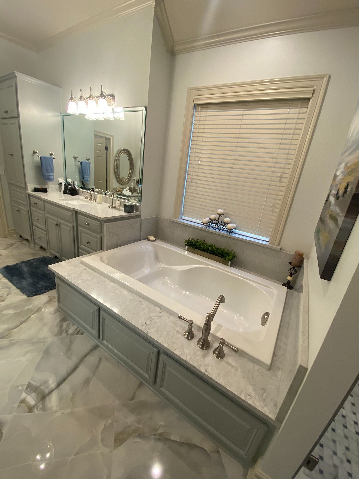 A bathroom featuring a white soaking tub with a marble surround, gray cabinets, a double vanity, and a window with blinds.