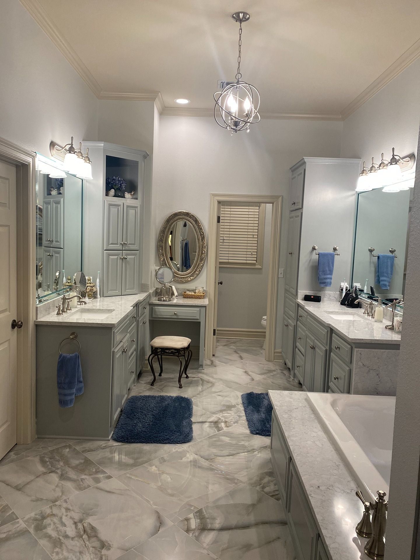 A spacious bathroom with gray cabinetry, marble-style flooring, a vanity, two mirrors, a light fixture, and a bathtub.