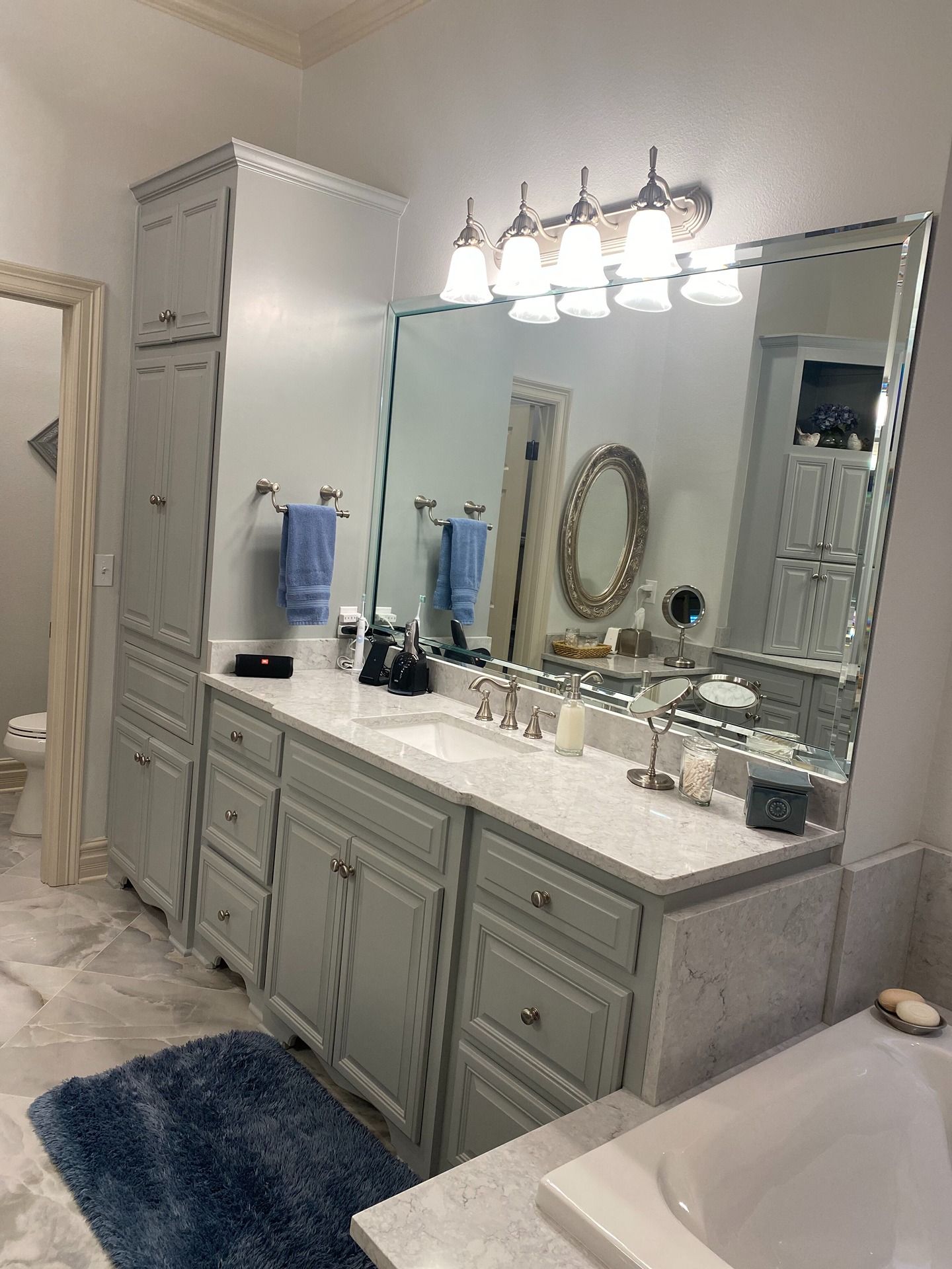A bathroom vanity with gray cabinets, a white countertop, large mirror, and overhead lights, next to a blue bath mat.