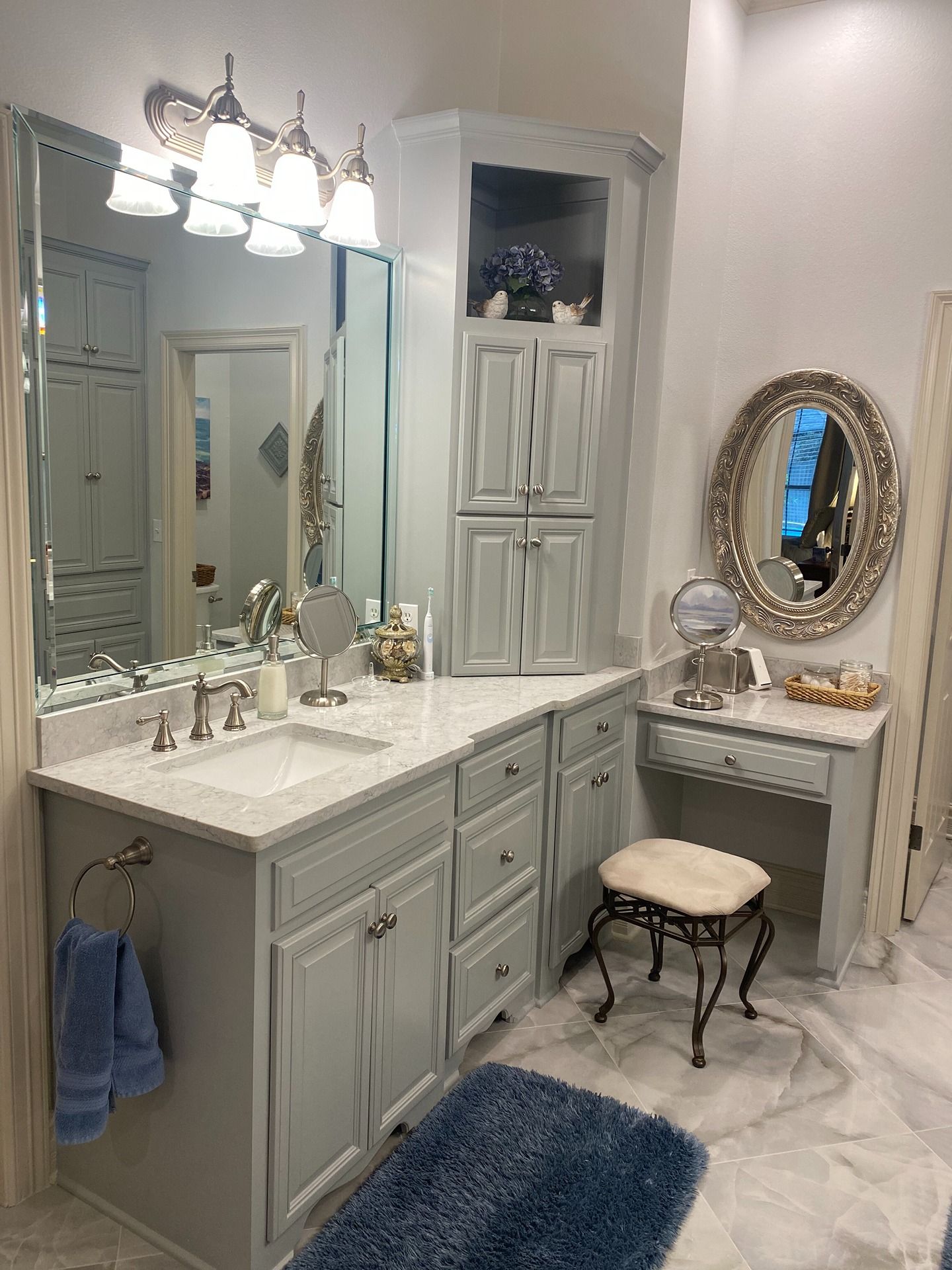 A bathroom vanity with gray cabinets, white marble countertop, tall corner storage, a large mirror, and a makeup station.
