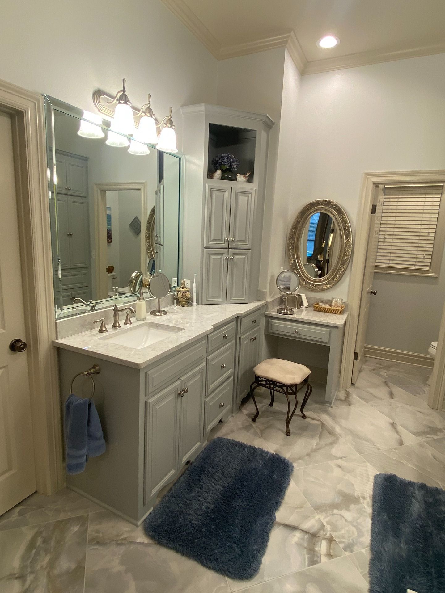 A gray bathroom vanity with a white countertop, mirror, corner cabinet, vanity stool, and blue rugs on tiled flooring.