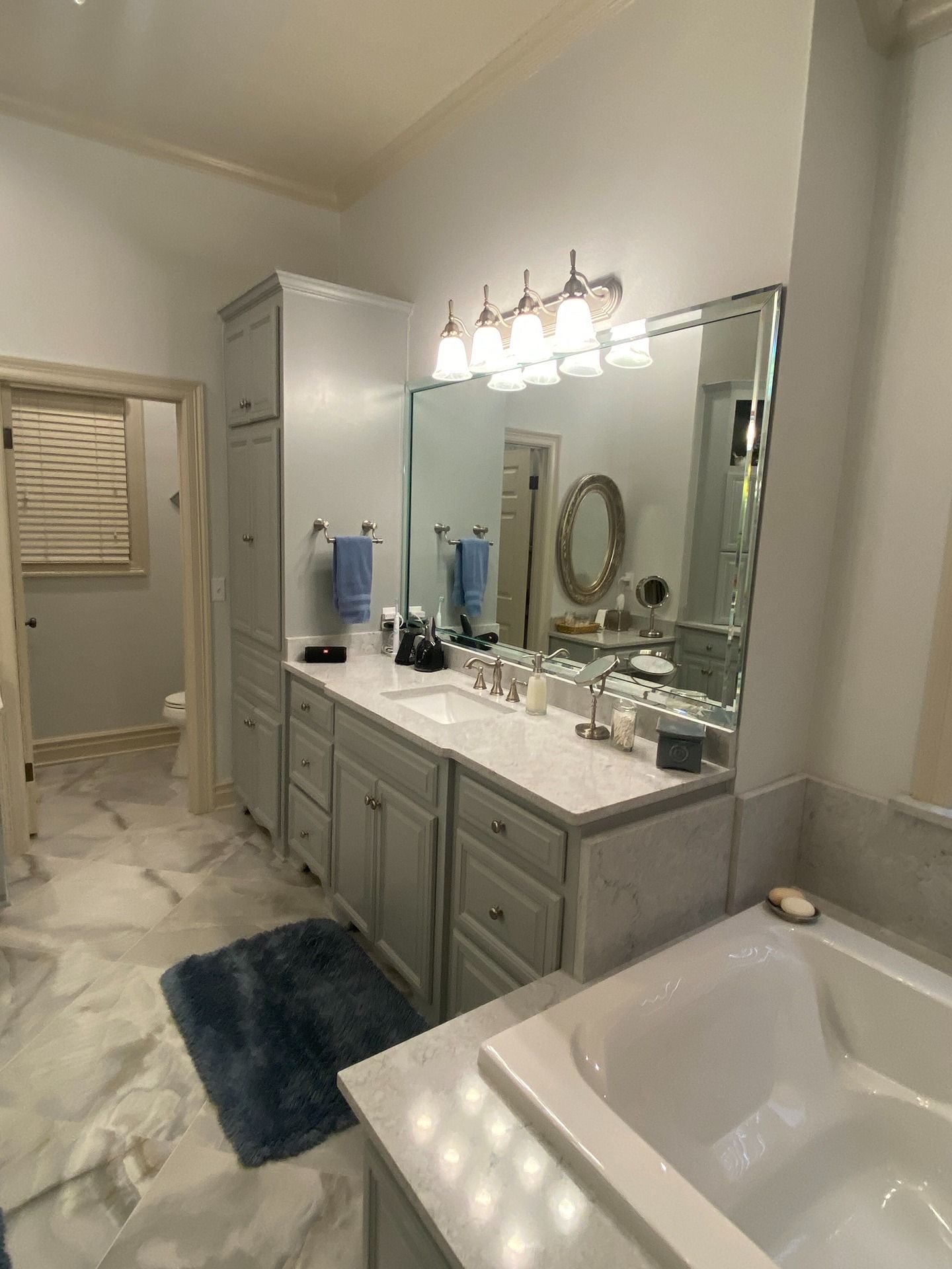 A bathroom with a gray vanity, white marble countertops, a large mirror, light fixtures, and a blue rug on tiled floors.