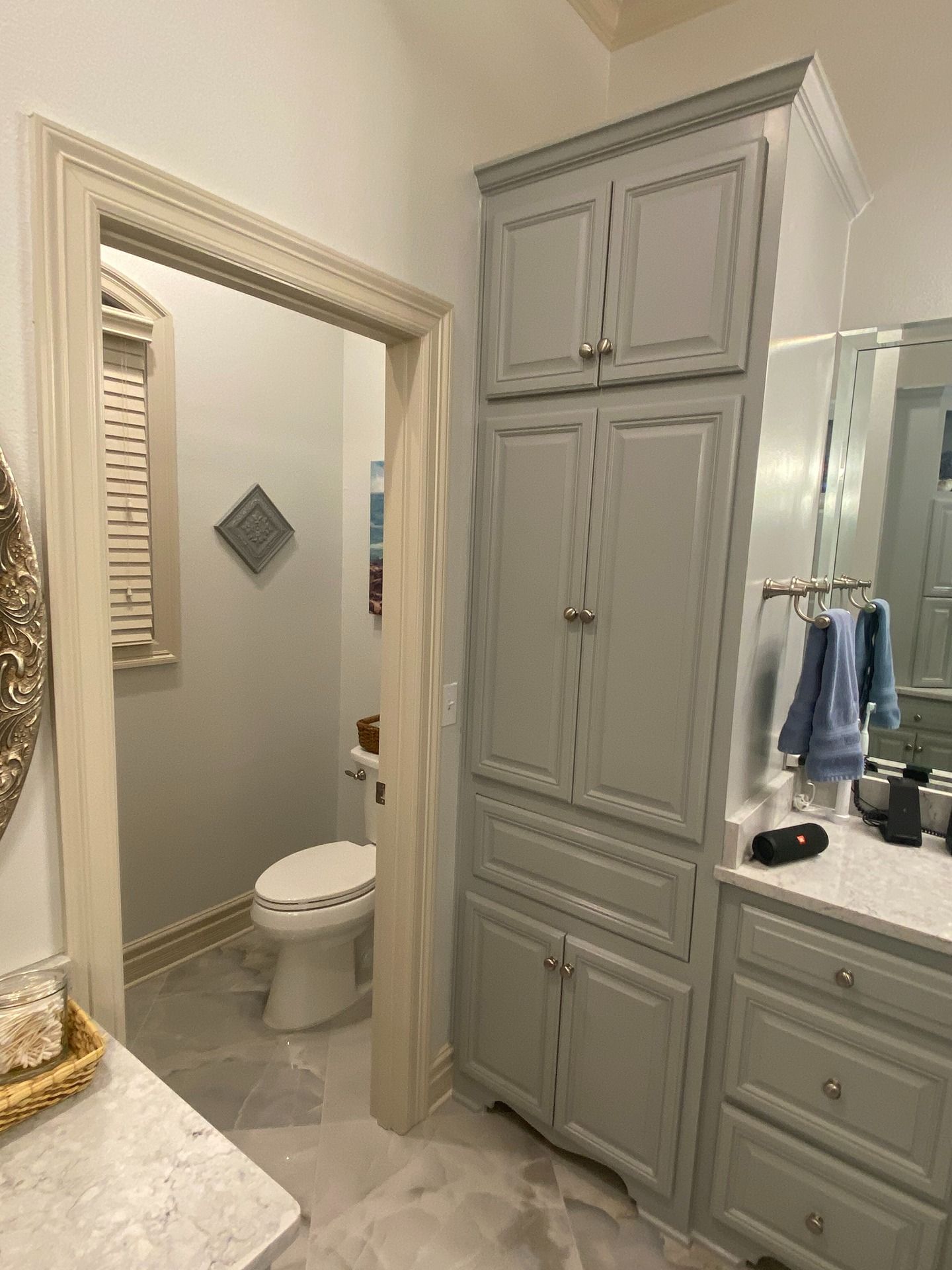 A gray linen cabinet stands in a bathroom next to a doorway leading to a toilet.