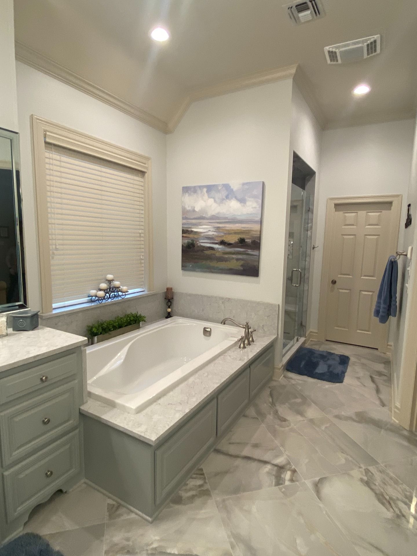A neutral-toned bathroom with a marble-tiled tub surround, a landscape painting, a gray vanity, and a glass-enclosed shower.