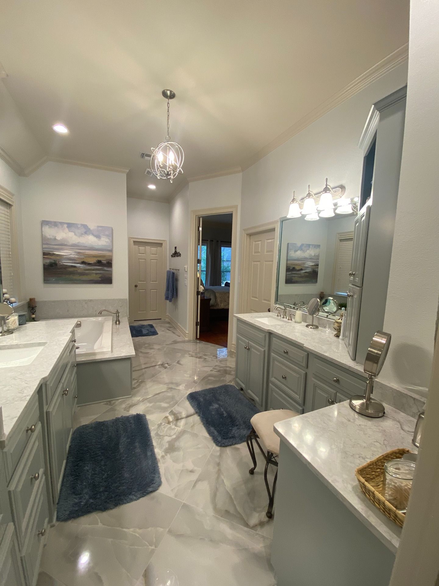 A spacious, brightly lit bathroom featuring marble-look floors, blue rugs, dual gray vanities, and a built-in bathtub.