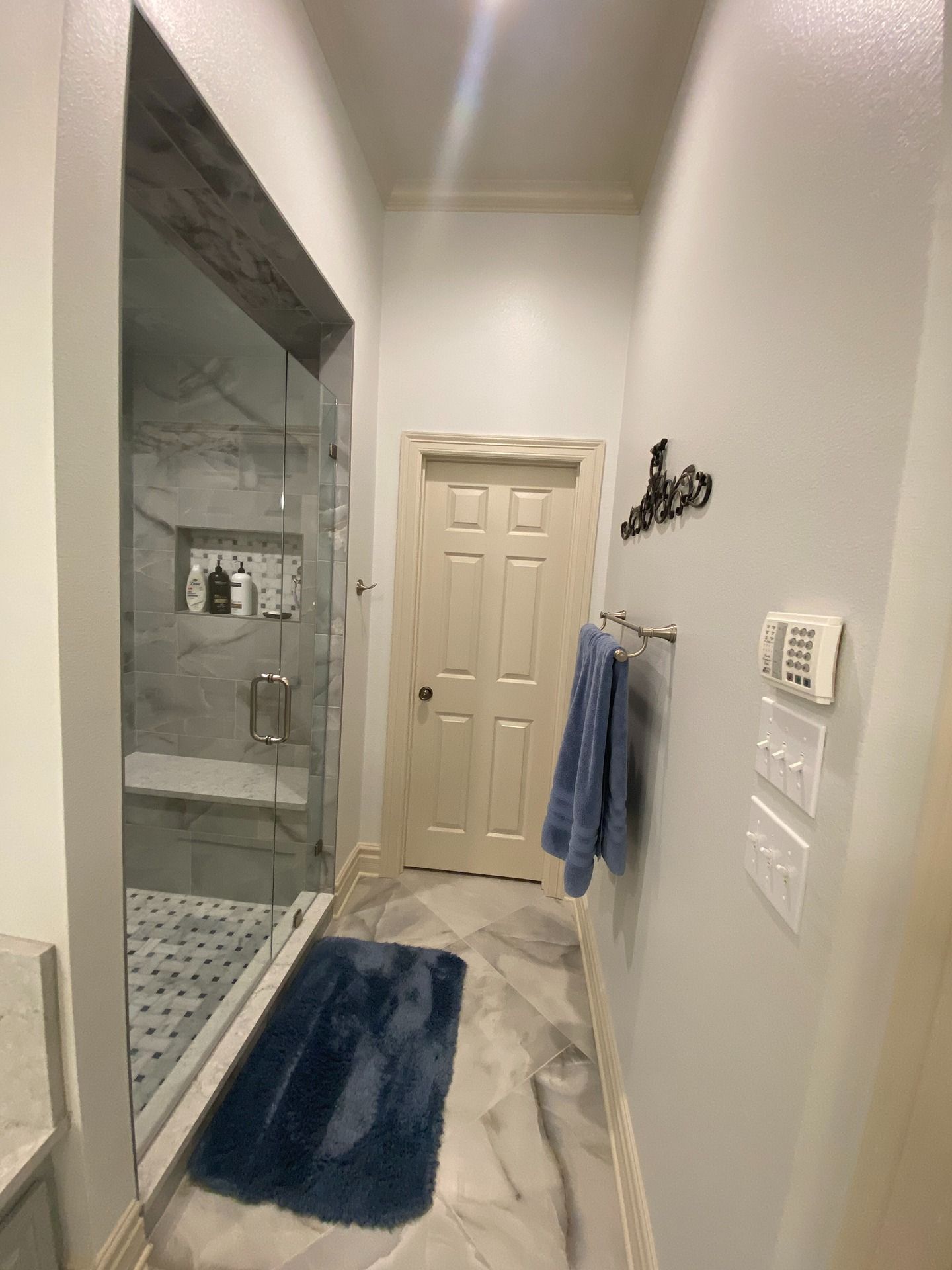 A bathroom features a glass-enclosed shower, a blue bath mat on white tile, and a closed door at the end of the hallway.