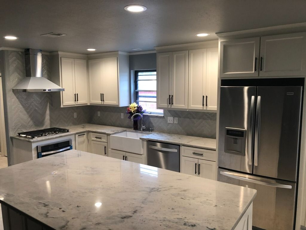 Modern kitchen with white cabinets, stainless steel appliances, a large marble island, and gray herringbone tile backsplash.