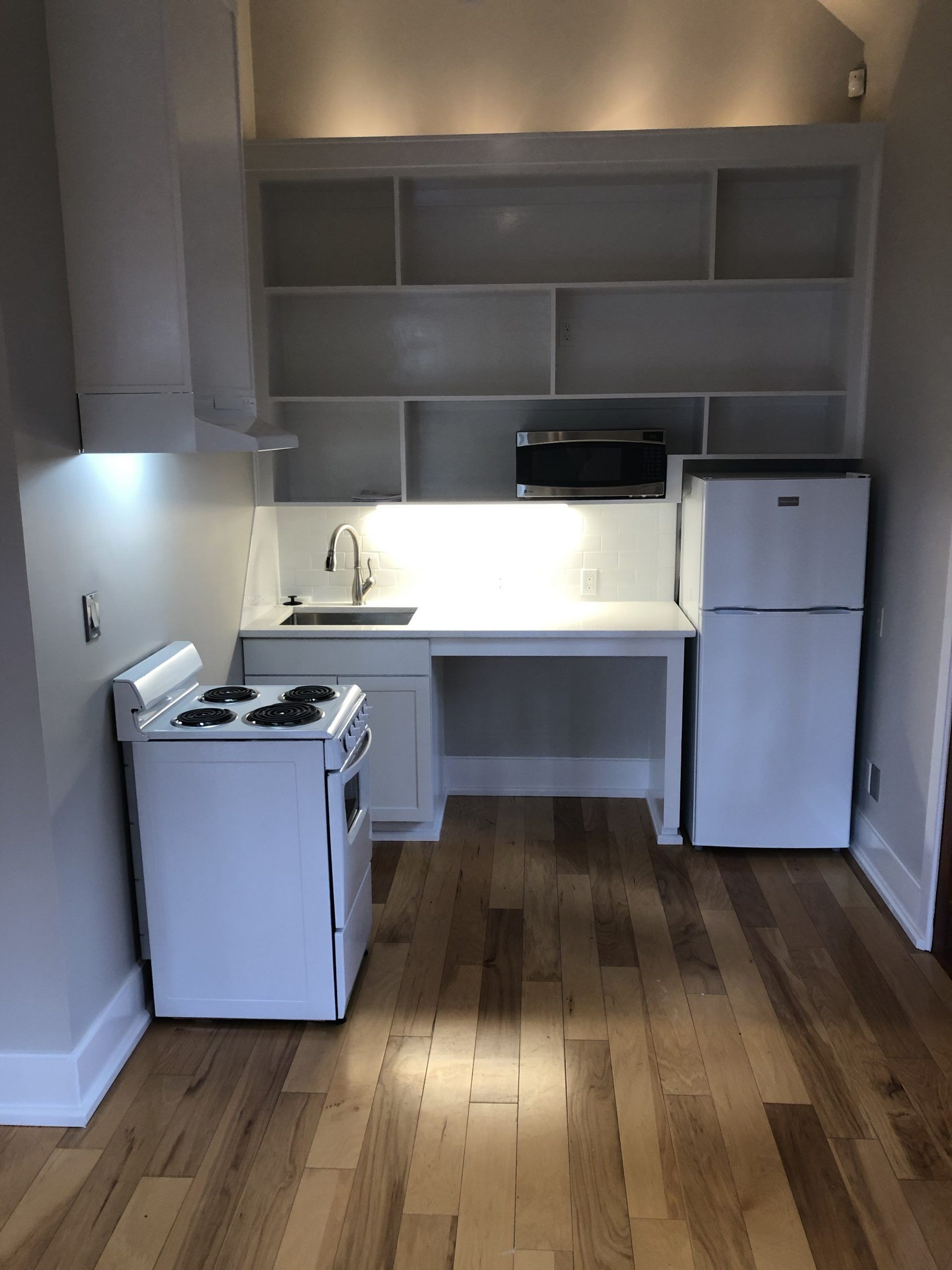 A small kitchenette with white appliances, a sink, open shelving, and light wood flooring.