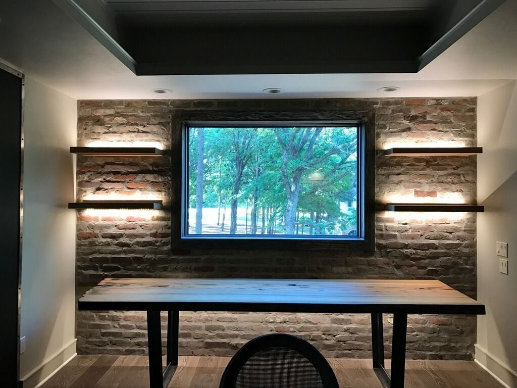 A wooden table sits before a rustic brick wall with four illuminated floating shelves and a central window viewing trees.