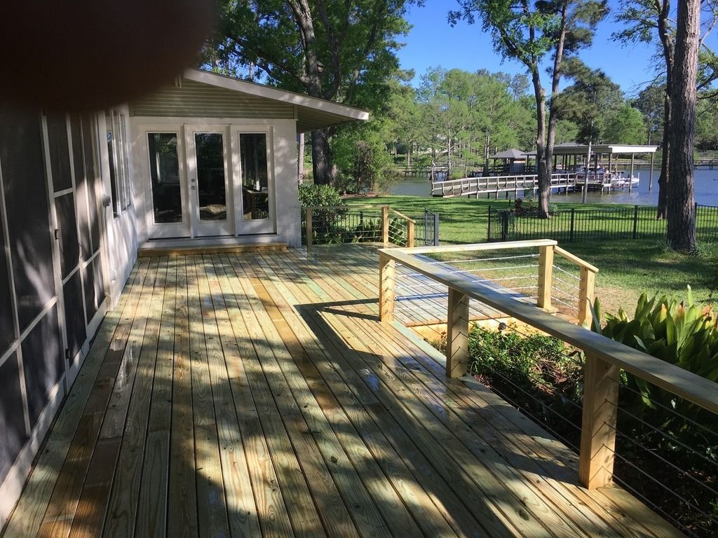 A newly constructed wooden deck with railings overlooks a grassy yard, trees, a dock, and a body of water under a blue sky.