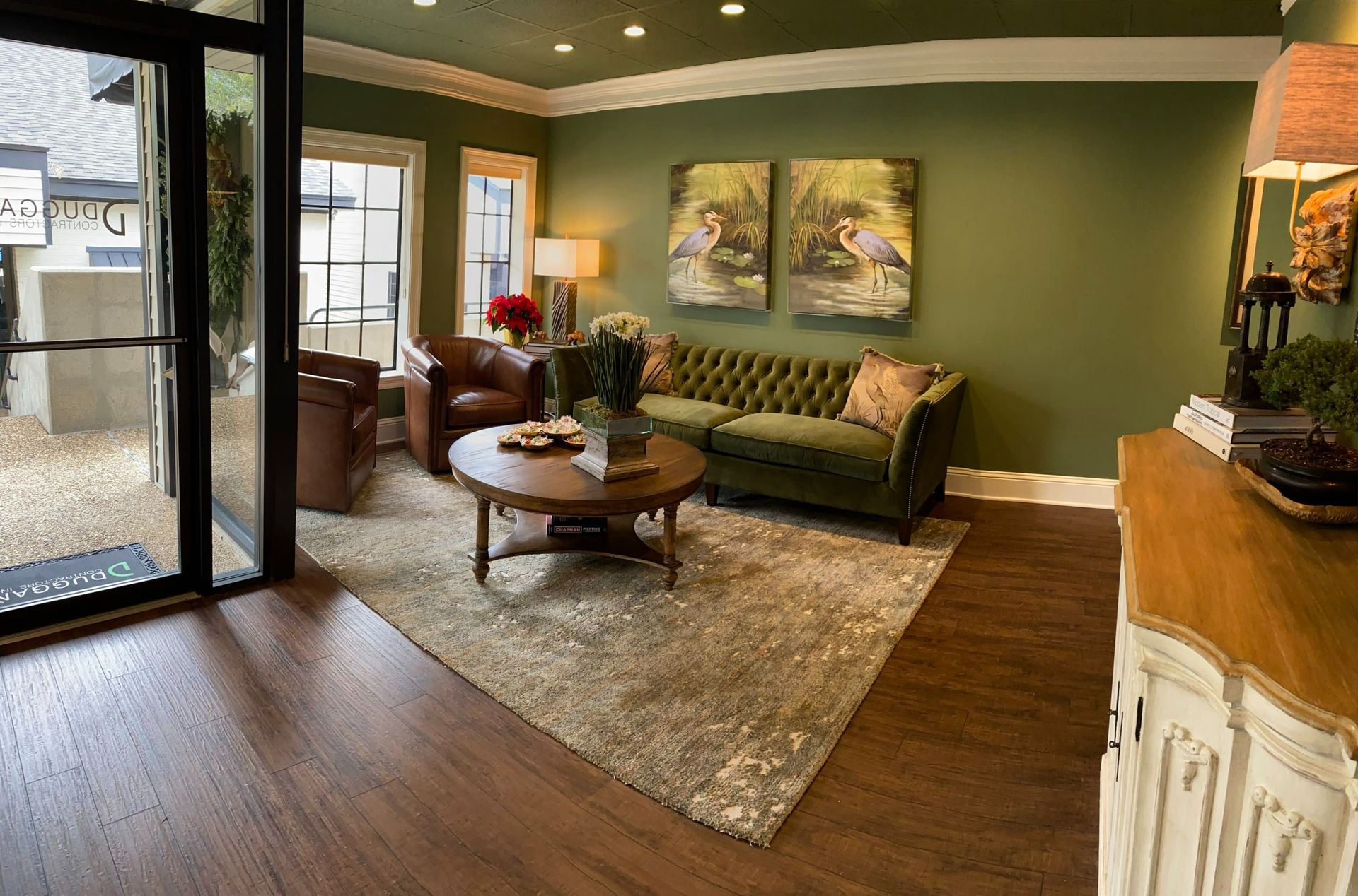 A cozy waiting room features a green tufted sofa, brown leather armchairs, a round wooden coffee table, and nature art.