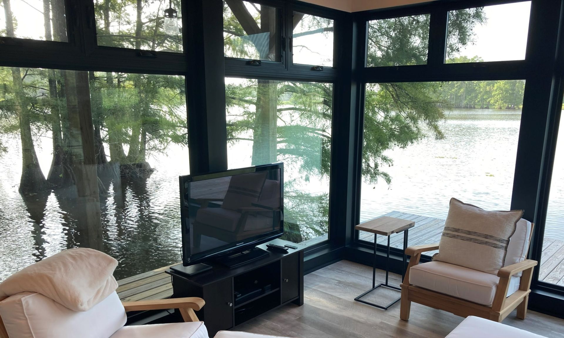 A cozy sunroom featuring large windows overlooking a lake, with two armchairs, a side table, and a television on a stand.