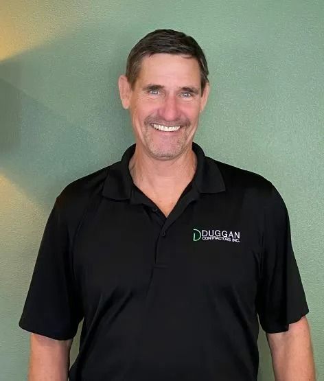 A smiling person wearing a black polo shirt with a company logo, standing against a textured green wall.