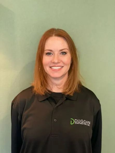 A person with reddish hair smiling against a solid green background, wearing a black long-sleeved Duggan Contracting shirt.