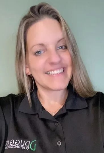A smiling professional wearing a black collared work shirt with the green Duggan Contractors logo, against a green wall.