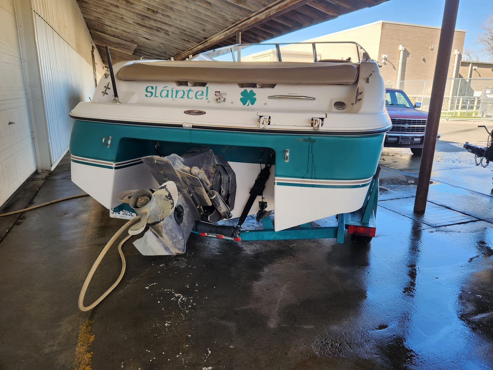 The rear view of a white and teal motorboat on a trailer under a covered area, with a clover decal and