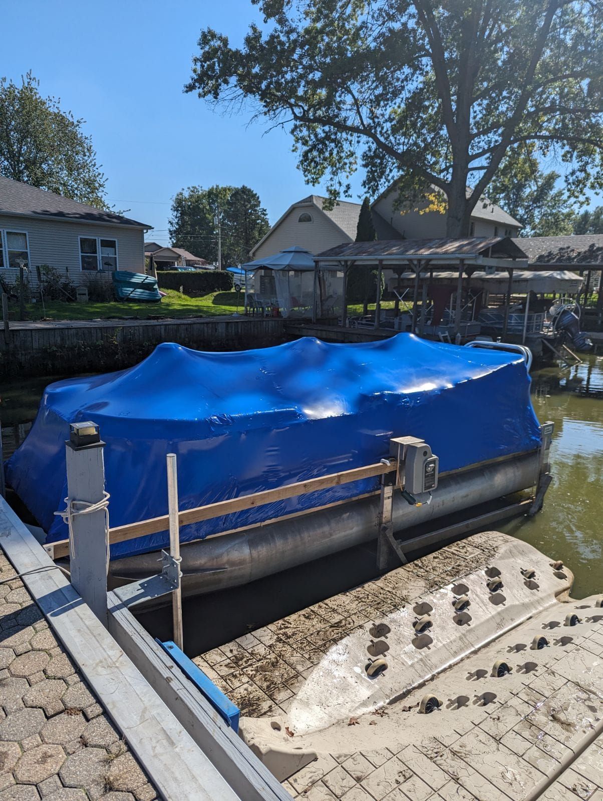 A pontoon boat with a bright blue cover parked at a dock next to a residential waterfront property on a sunny day.