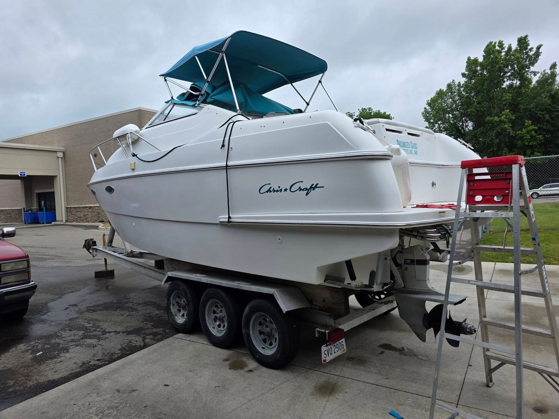 A white Chris-Craft boat on a triple-axle trailer with a teal canopy, parked on a paved lot beside a red ladder.