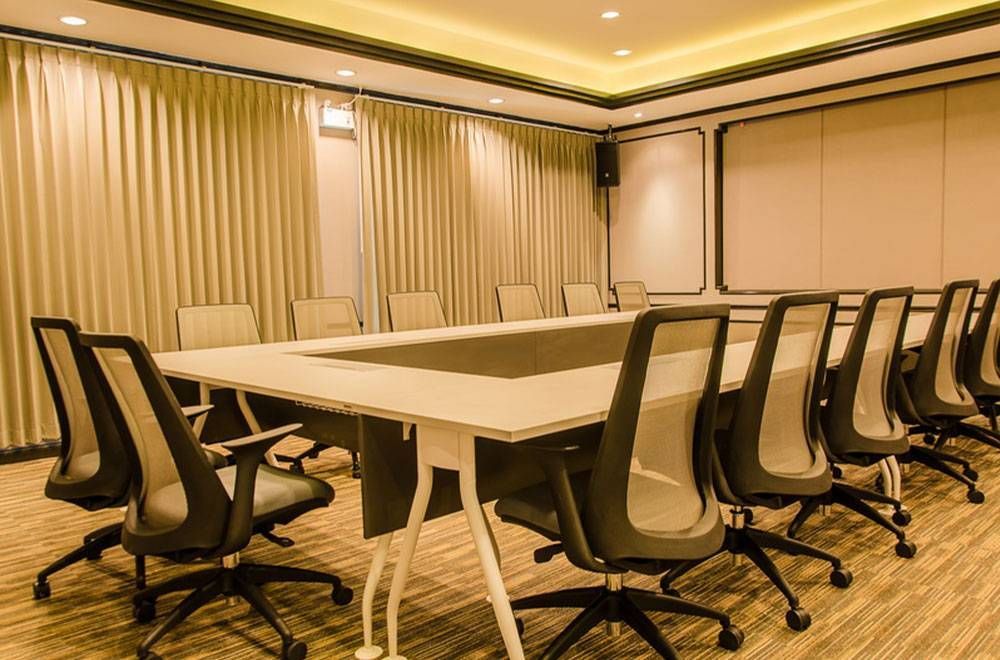 ห้องประชุมที่มีโต๊ะสี่เหลี่ยมผืนผ้าและเก้าอี้สำนักงาน ม่านสีเบจ และแสงไฟโทนอบอุ่น