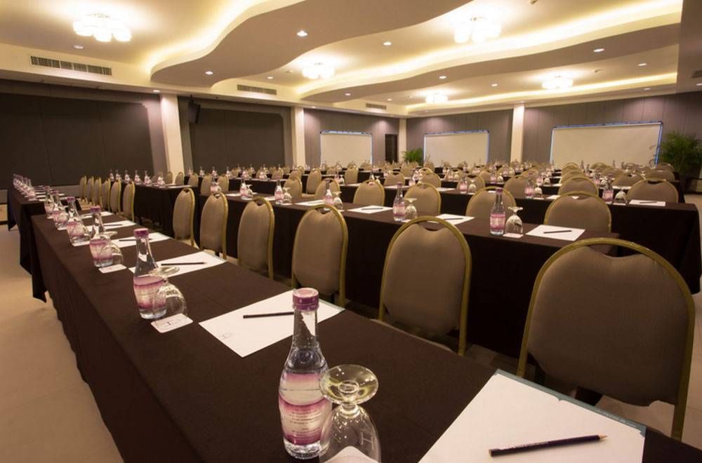 ห้องประชุมจัดเตรียมด้วยโต๊ะเรียงเป็นแถว ผ้าปูโต๊ะสีน้ำตาล ขวดน้ำ และเก้าอี้