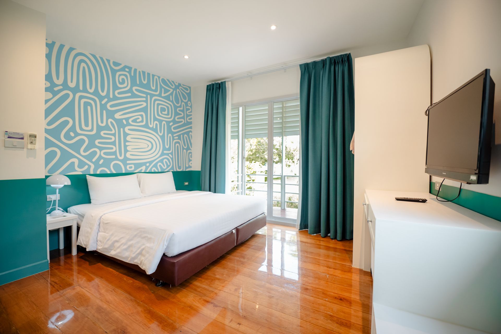 Bedroom with a bed, teal accents, patterned wall, and a TV. Sunlight streams through a window.