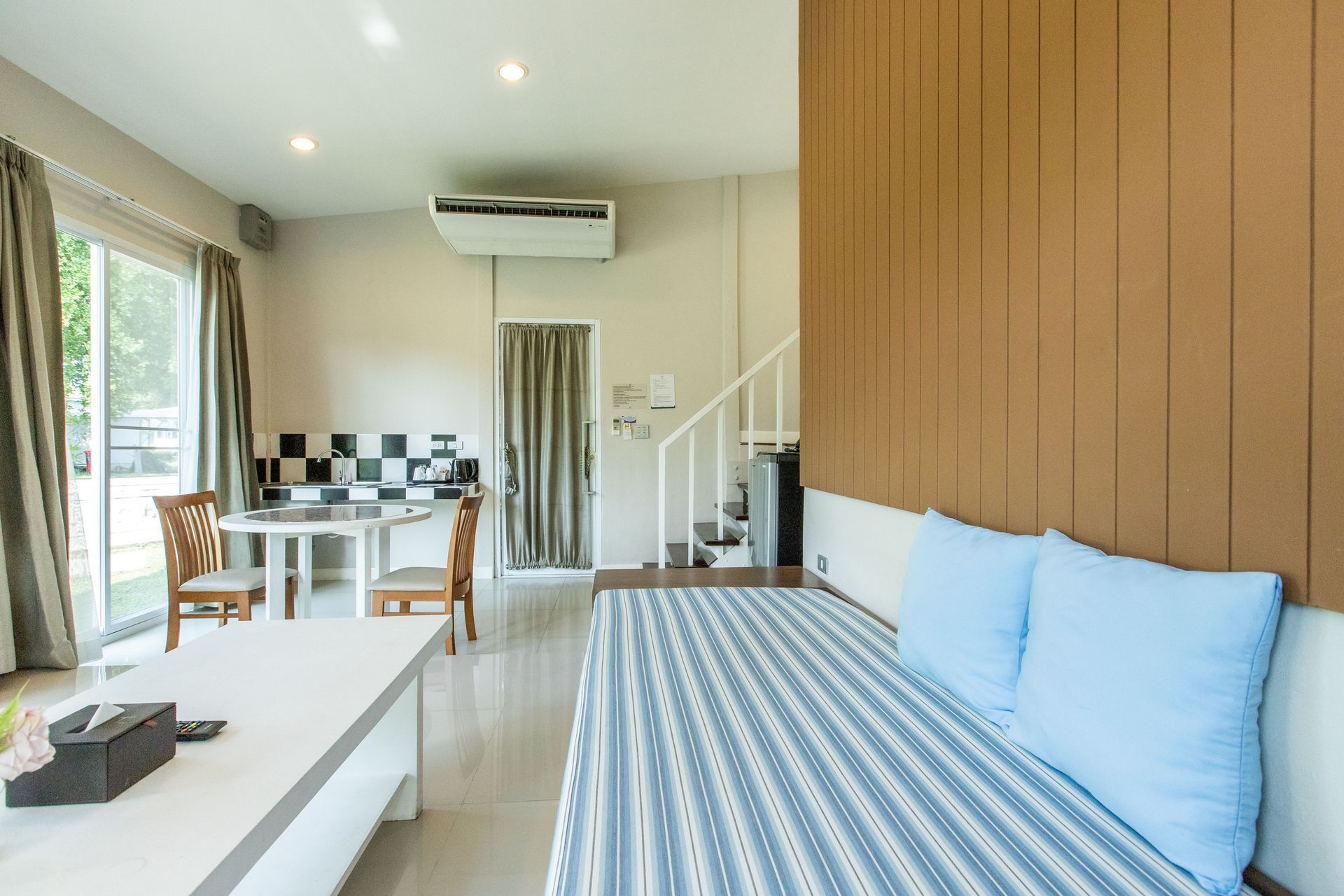 ภายในห้องนั่งเล่นขนาดเล็กที่มีห้องครัว พื้นที่รับประทานอาหาร และโซฟา ผนังและบันไดตกแต่งด้วยแผ่นไม้