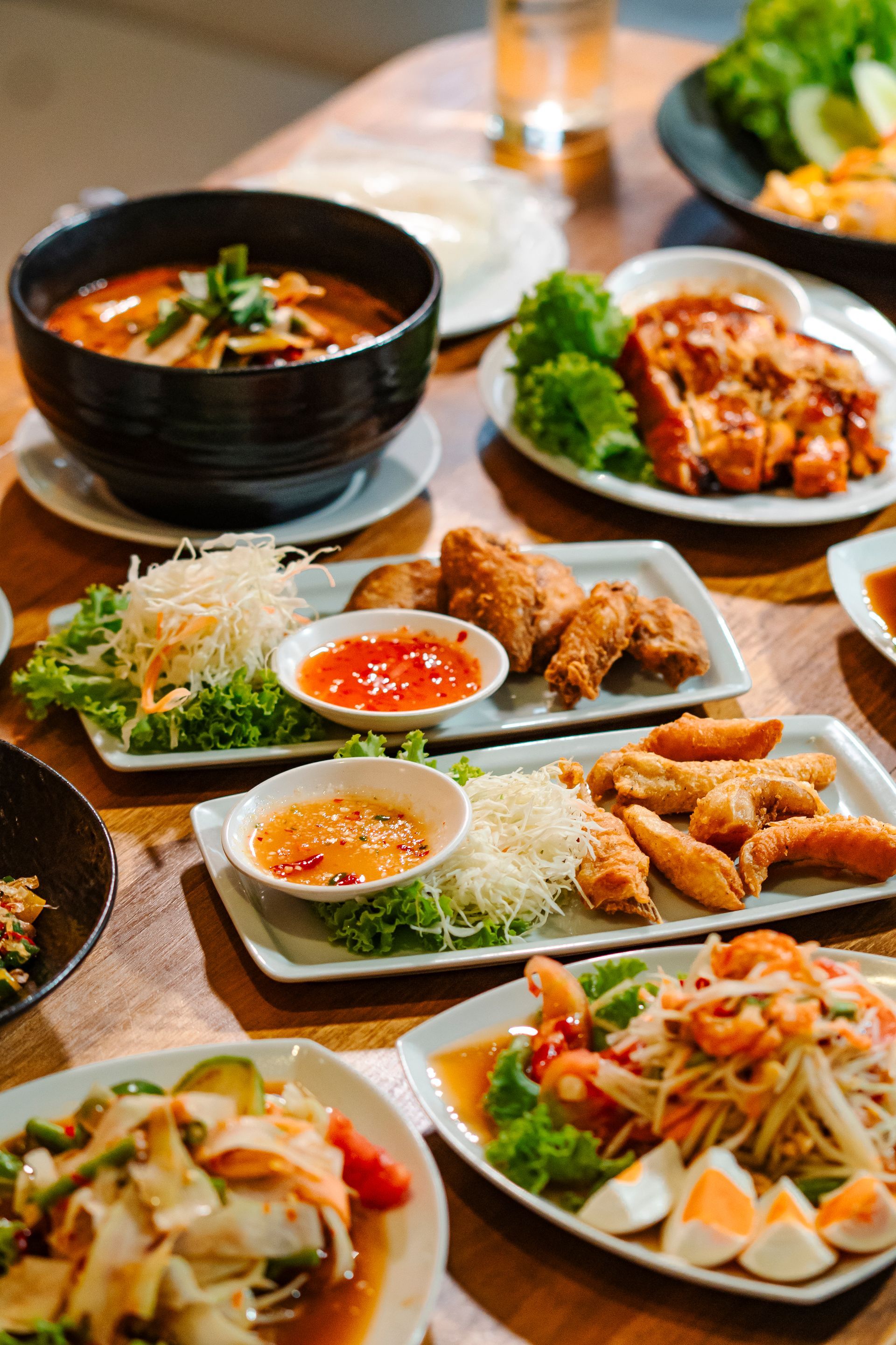 อาหารไทยหลากหลายชนิดจัดวางอยู่บนโต๊ะไม้ ได้แก่ ซุป ส้มตำ ไก่ทอด ปอเปี๊ยะ และน้ำจิ้ม