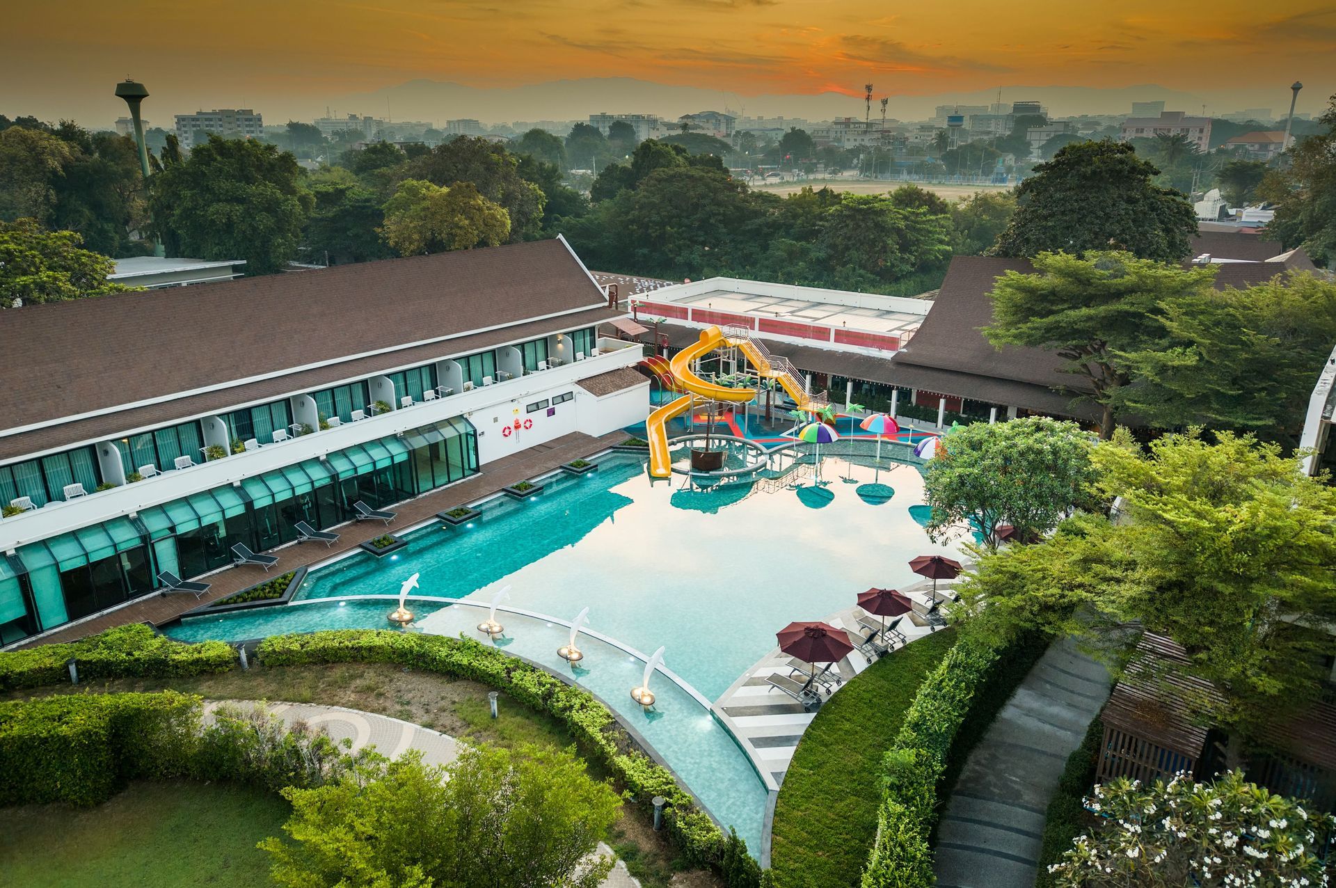 ภาพถ่ายทางอากาศของรีสอร์ทที่มีสระว่ายน้ำ สไลเดอร์น้ำ และอาคารต่างๆ ล้อมรอบด้วยต้นไม้เขียวขจี ท้องฟ้ามีสีสันยามพระอาทิตย์ตกดิน