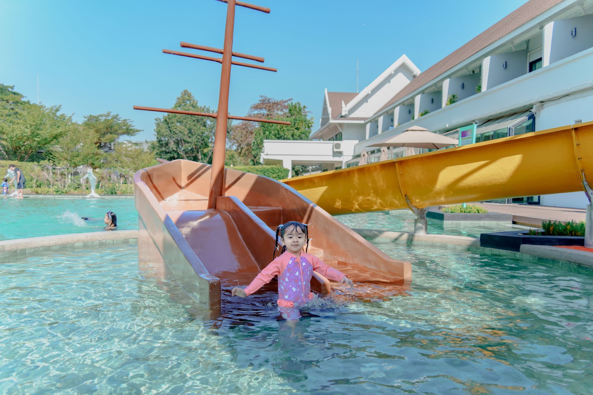 หญิงสาวในชุดว่ายน้ำกำลังออกจากสไลเดอร์น้ำรูปเรือลงสู่สระว่ายน้ำ ในวันที่มีแดดจัด โดยมีโรงแรมอยู่ด้านหลัง