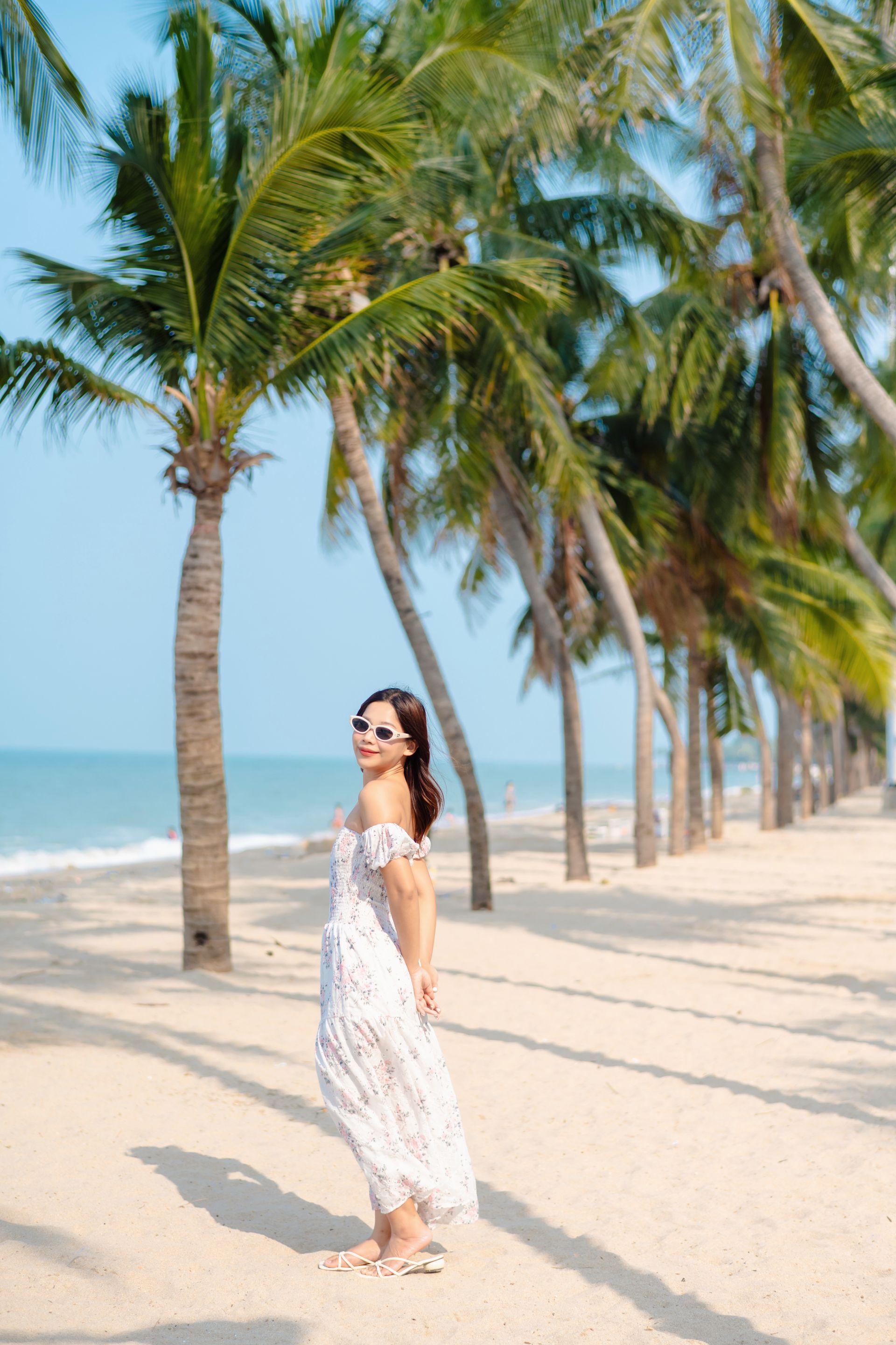 หญิงสาวในชุดเดรสสีขาวและแว่นกันแดด บนหาดทราย โดยมีต้นปาล์มเป็นฉากหลัง