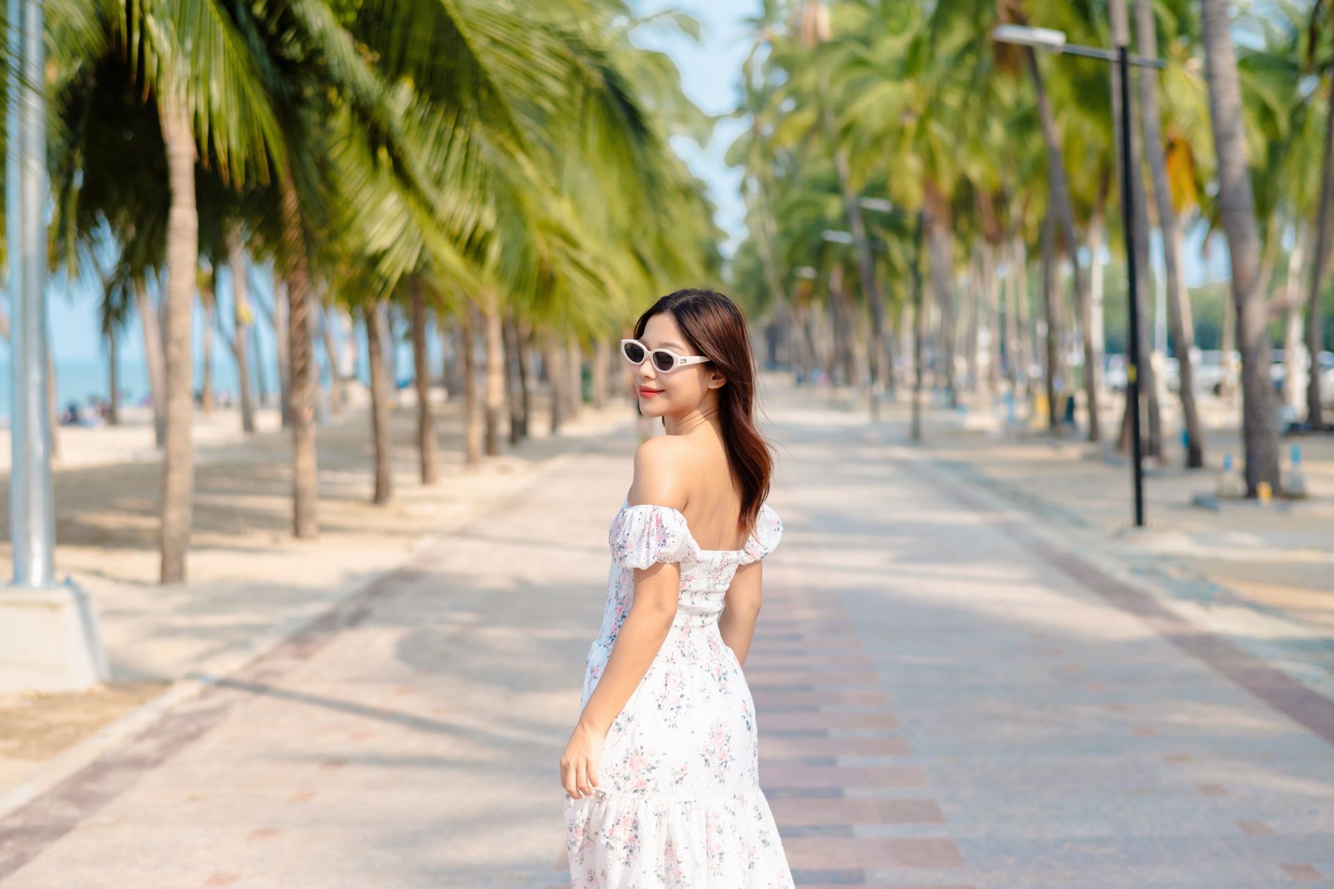หญิงสาวในชุดเดรสสีขาวและแว่นกันแดด ยิ้มพลางมองข้ามไหล่ไปยังทางเดินที่เรียงรายไปด้วยต้นปาล์ม