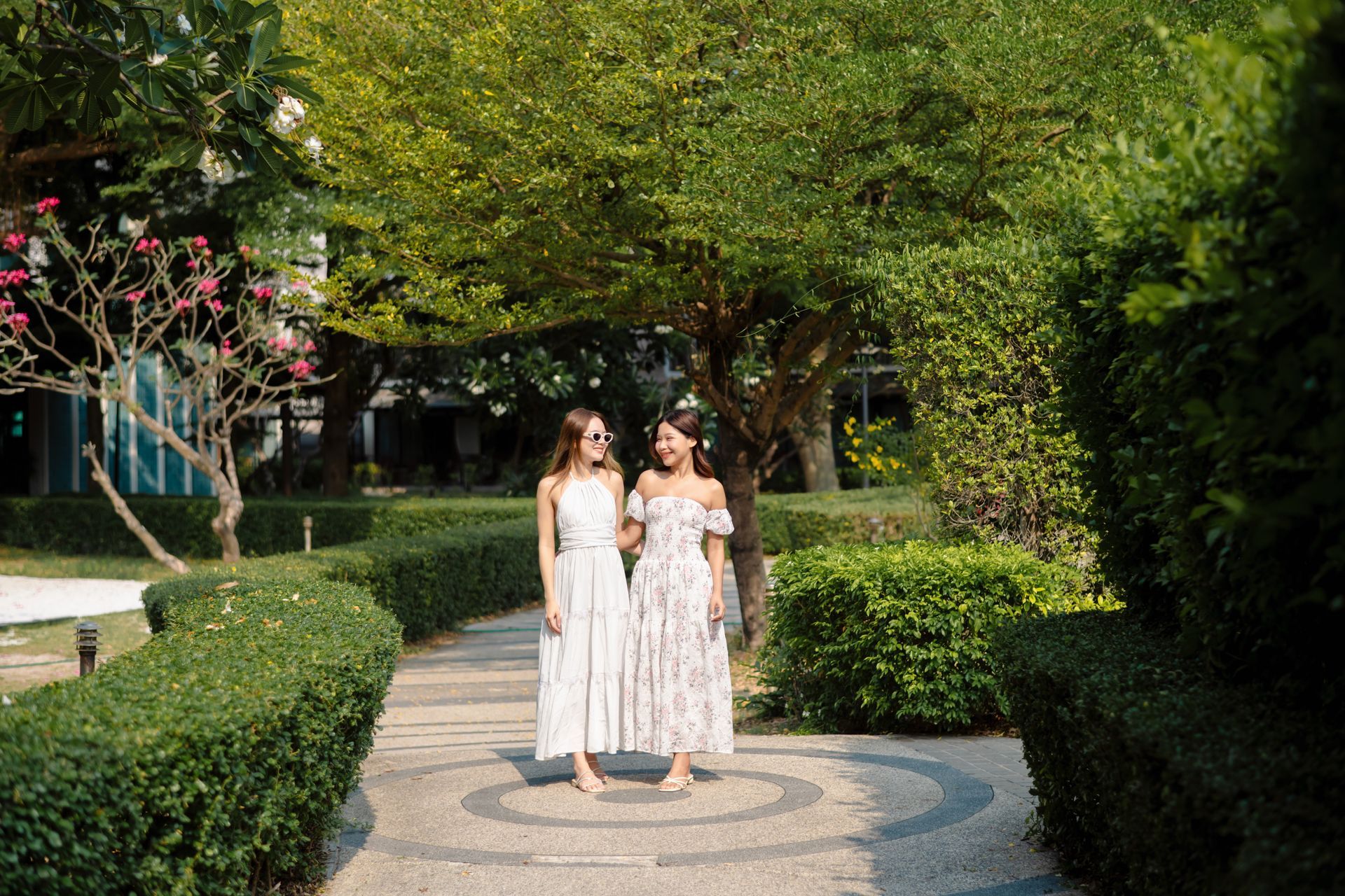 หญิงสาวสองคนในชุดเดรสสีขาวเดินอยู่บนทางเดินในสวนใต้ต้นไม้