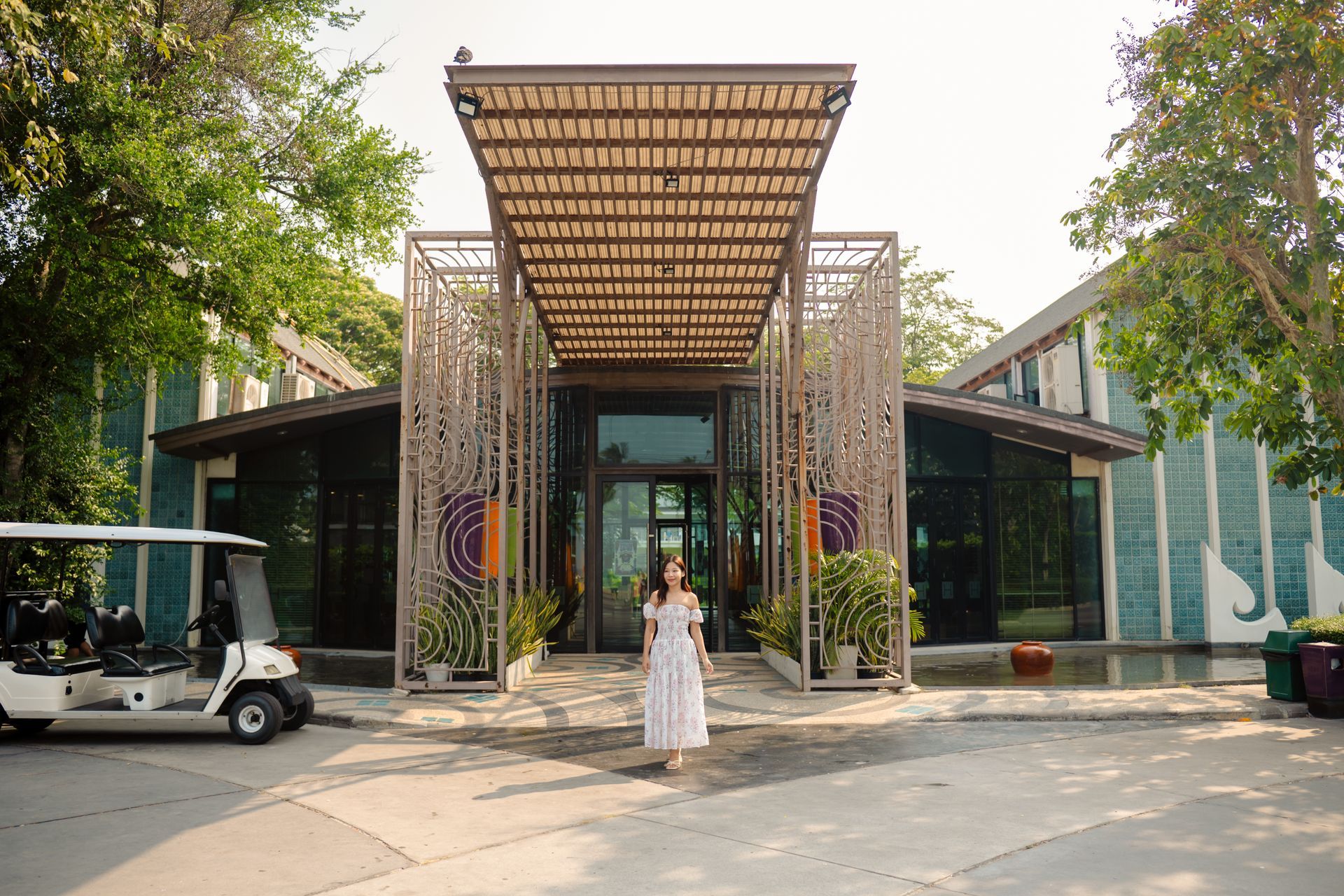 หญิงสาวสวมชุดเดรสยืนอยู่หน้าอาคาร โดยมีรถกอล์ฟอยู่ทางด้านซ้าย