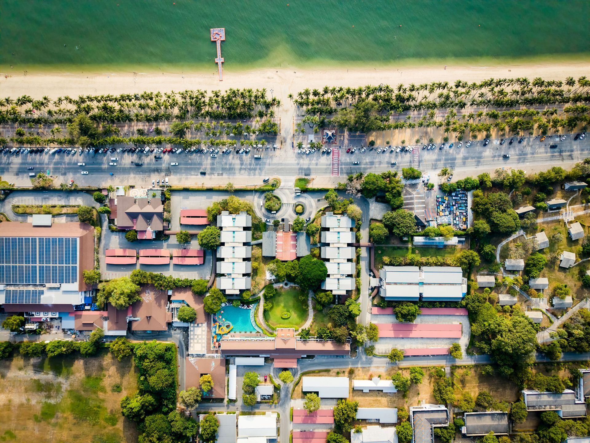 ภาพถ่ายทางอากาศของเมืองชายหาดที่มีหาดทราย ถนน อาคาร ต้นไม้ และท่าเรือ น้ำทะเลสีฟ้าและพืชพรรณสีเขียว