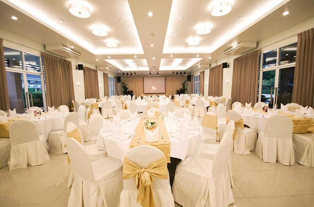 ห้องจัดเลี้ยงถูกจัดเตรียมไว้สำหรับจัดงาน มีโต๊ะกลม เก้าอี้สีขาวผูกริบบิ้นสีทอง ม่านสีเบจ และเวทีพร้อมจอฉายโปรเจ็กเตอร์