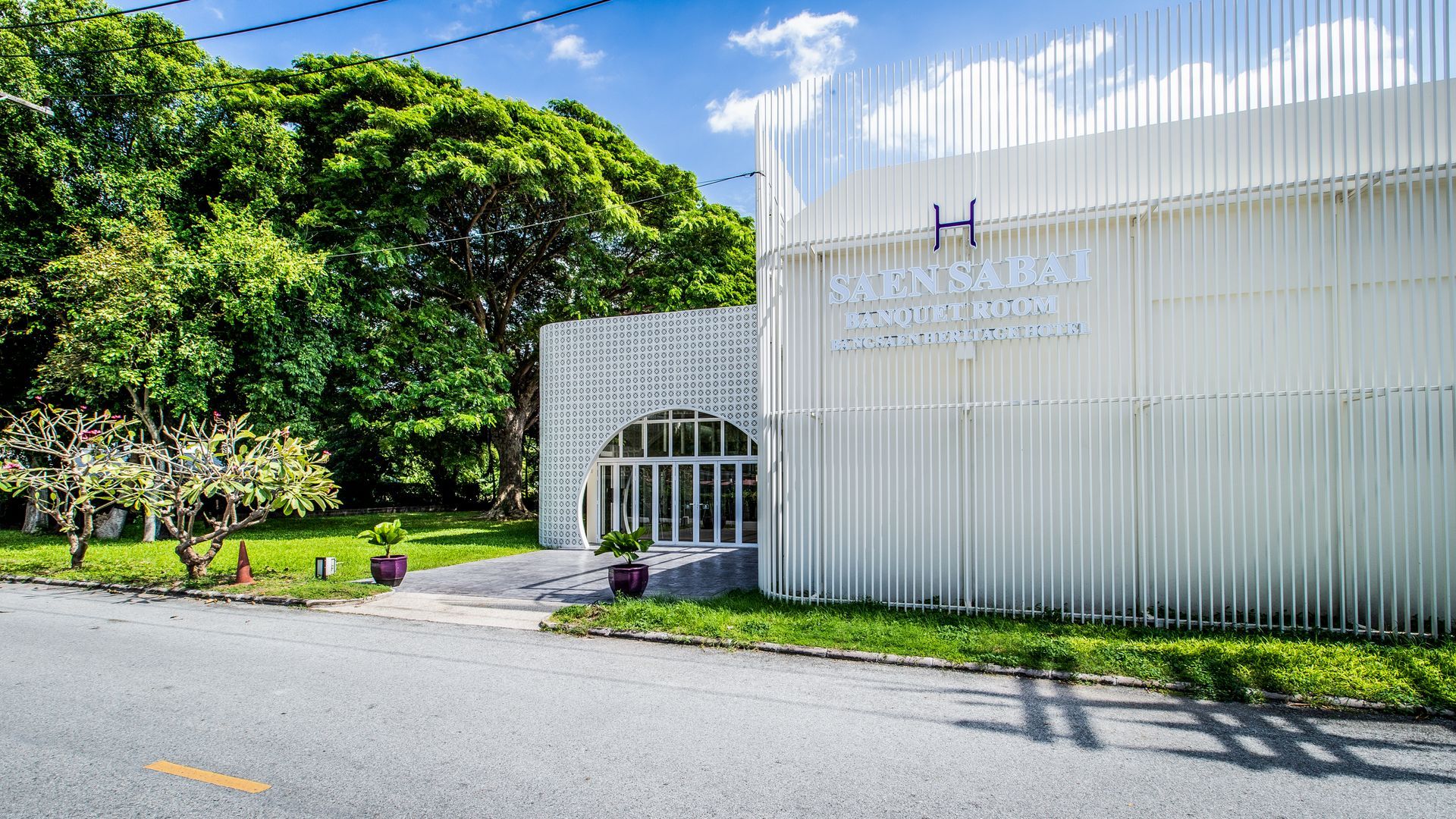 White building with vertical bars, arched entrance, and a sign partially visible. Green trees and grass surround.