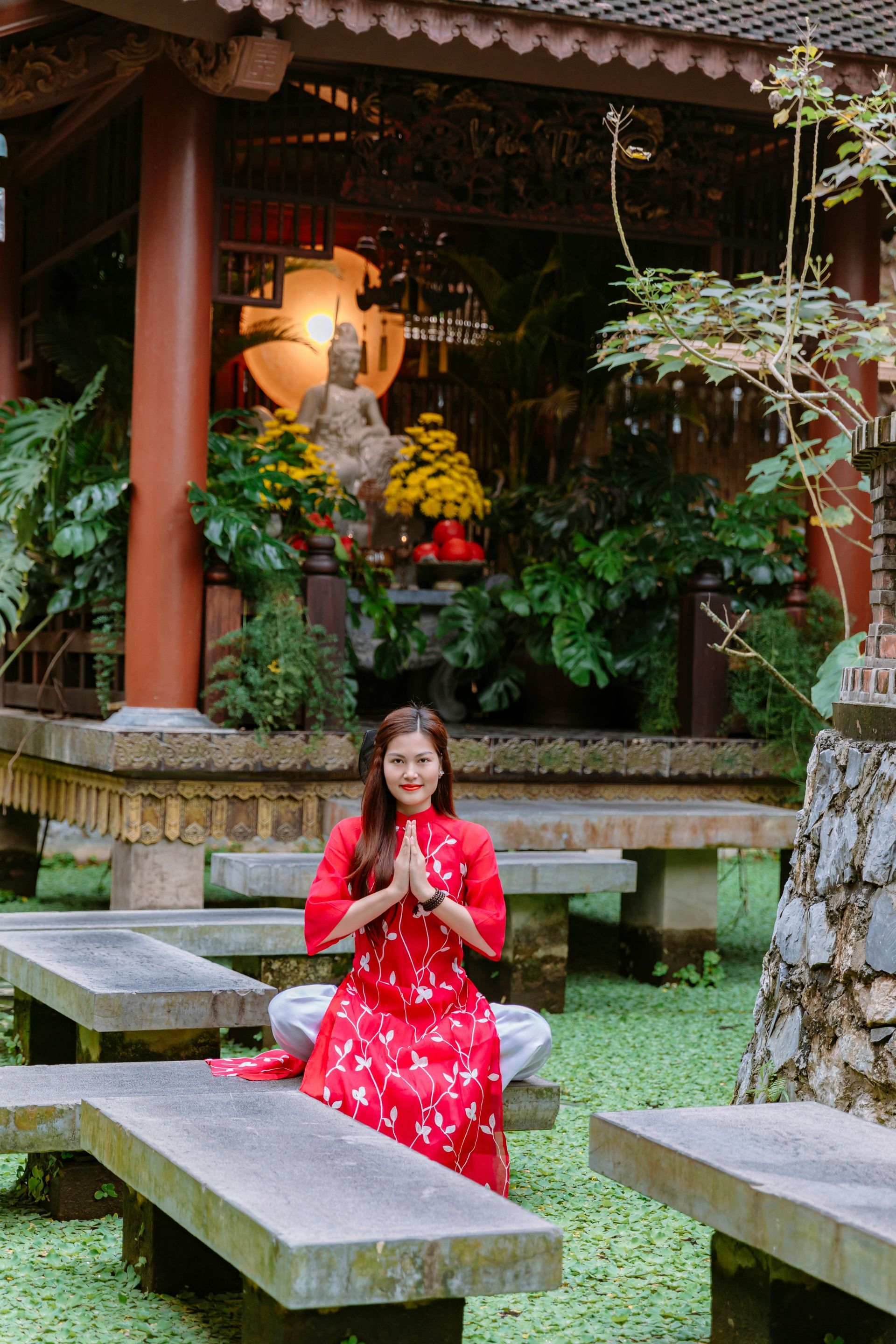 หญิงสาวในชุดสีแดง ยืนพนมมืออยู่หน้าวัดที่มีต้นไม้เขียวชอุ่ม