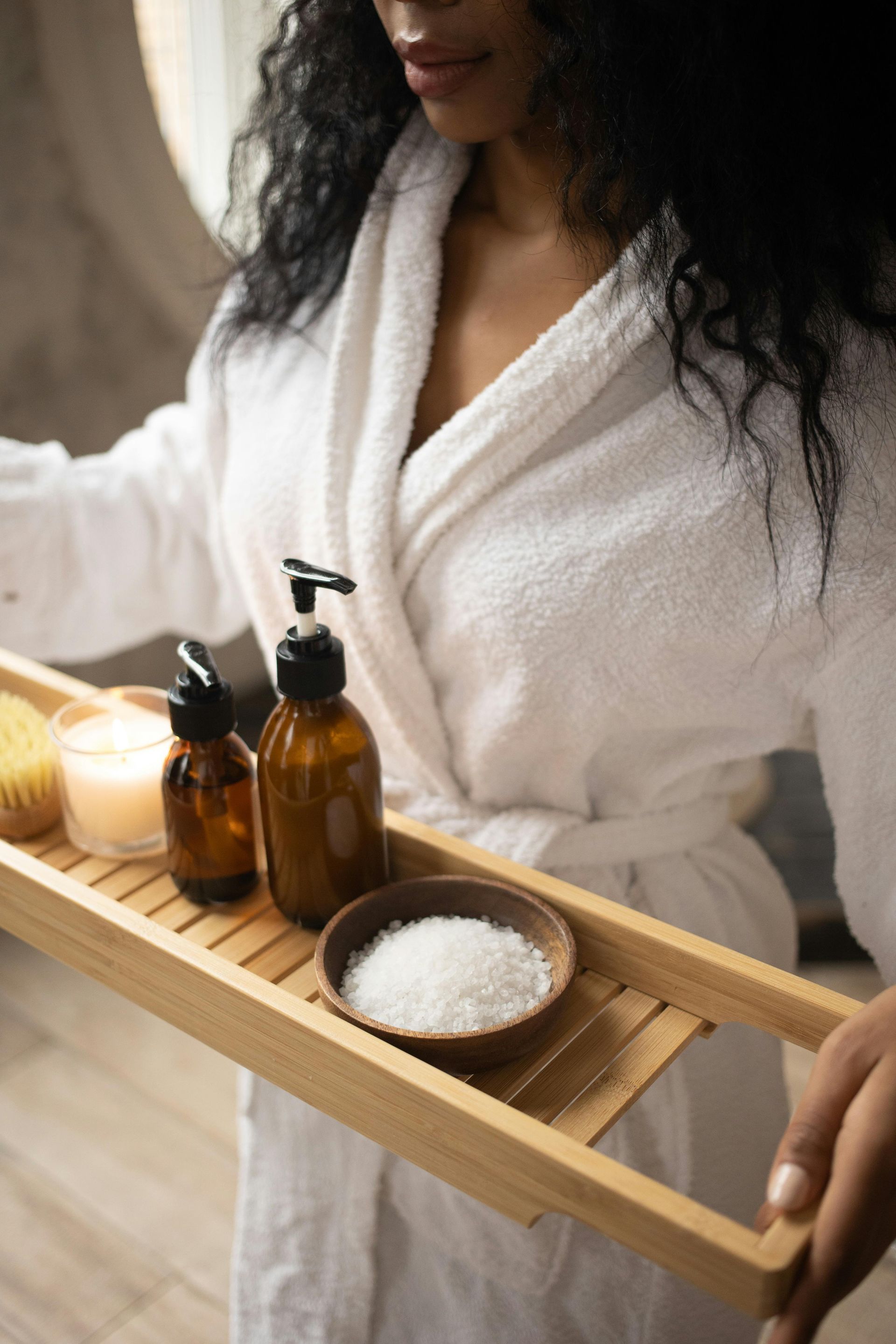 Woman in white robe holding tray with spa products: candle, oil bottles, and bath salts.