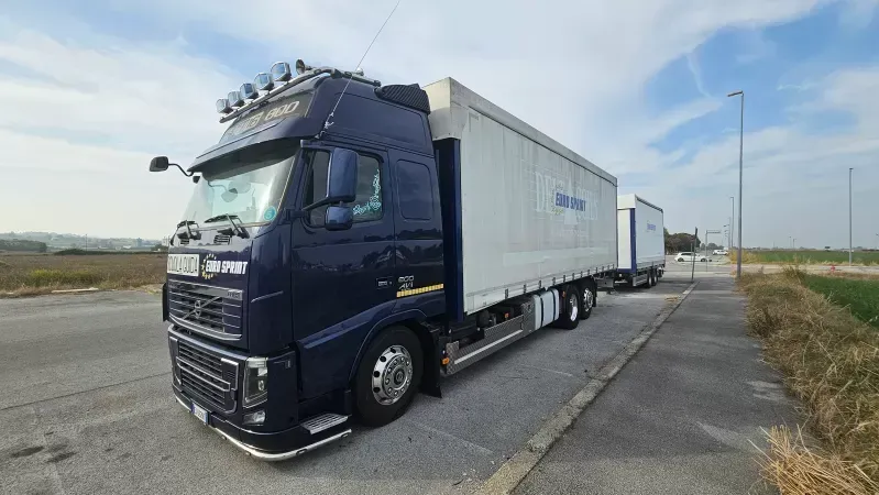 Un camion Volvo blu scuro con rimorchio bianco parcheggiato a lato della strada sotto un cielo azzurro brillante.