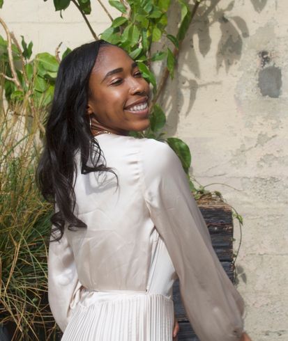 Smiling woman in a white dress looking over her shoulder beside green plants and a stucco wall