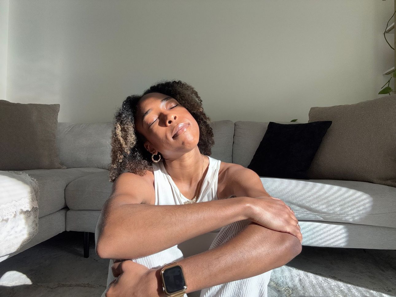 Person sitting on a couch with knees up, wearing a white tank top, head tilted, in a bright living room.