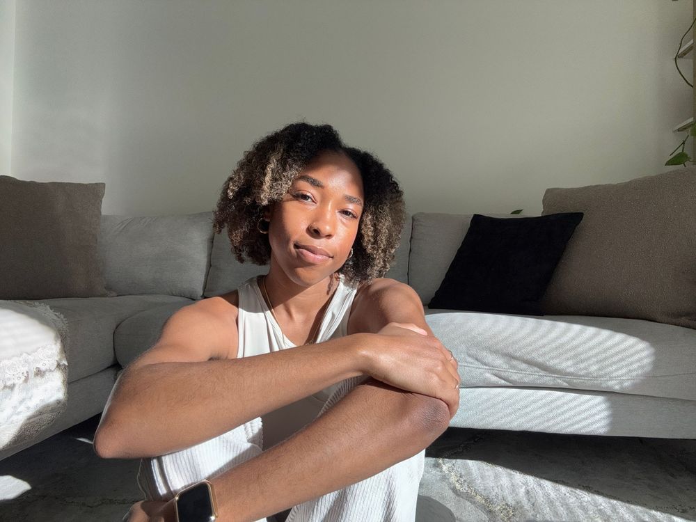 Person sitting on a sunlit living room floor beside a gray sofa, resting arms on knees and looking at the camera.